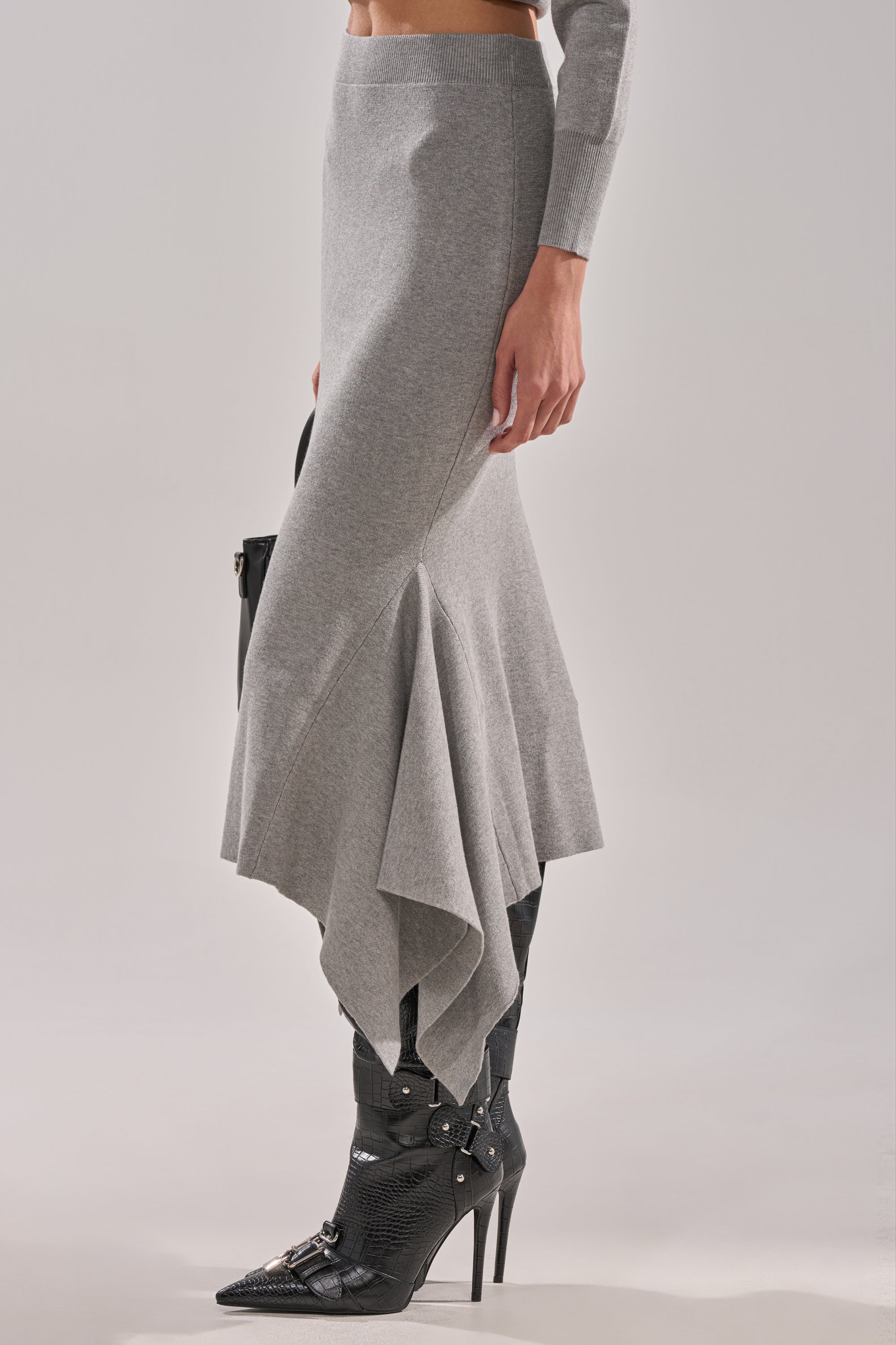 A person wears the LEXI MIDI SKIRT in gray—a stretchy knit midi skirt with an asymmetrical handkerchief hem—paired with black pointed-toe heeled boots, a long-sleeve top, and holds a black bag against a plain light background. 2