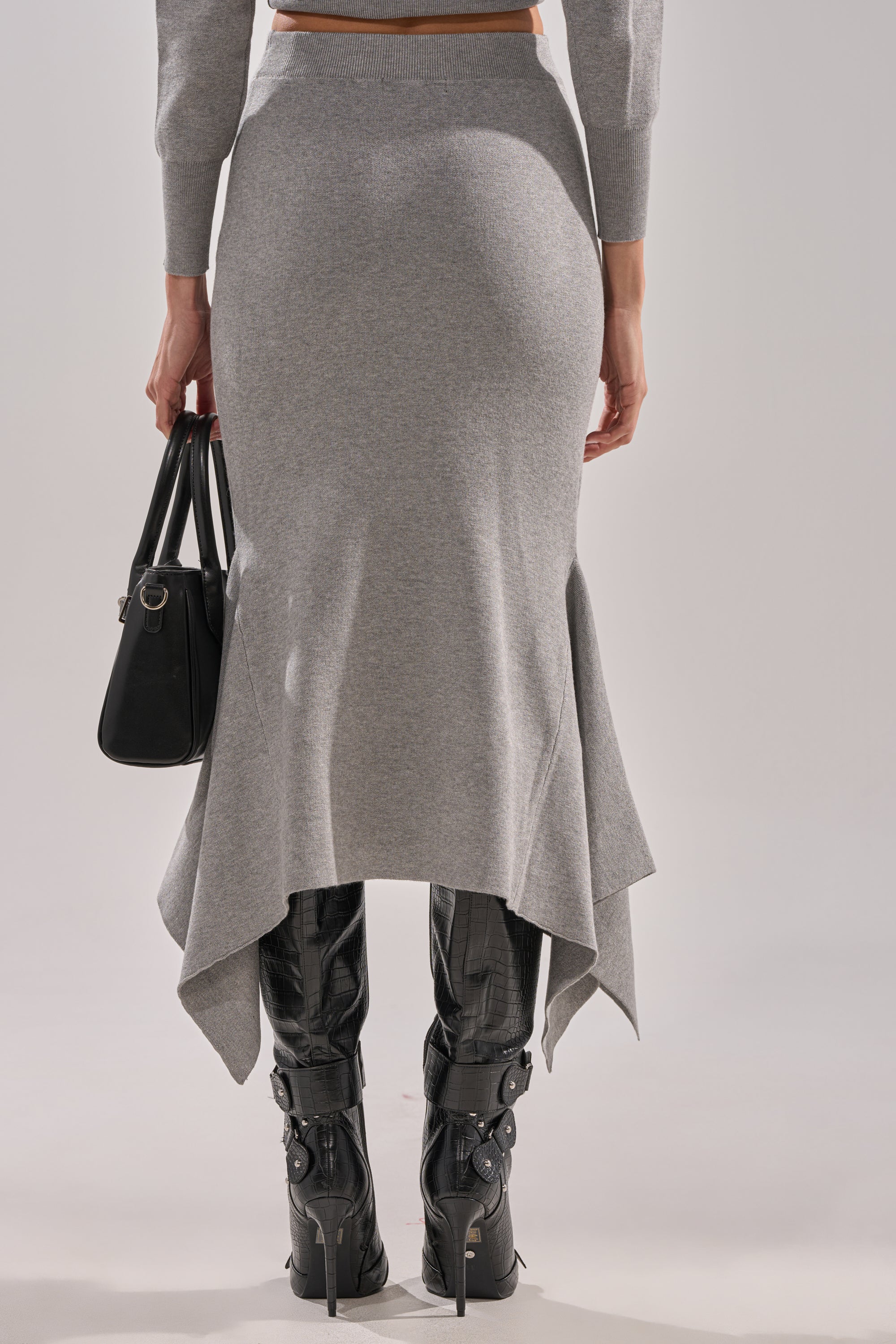 A person wearing the LEXI MIDI SKIRT with an asymmetrical hem, paired with black knee-high boots and a black handbag, is shown from the back against a plain background. 3