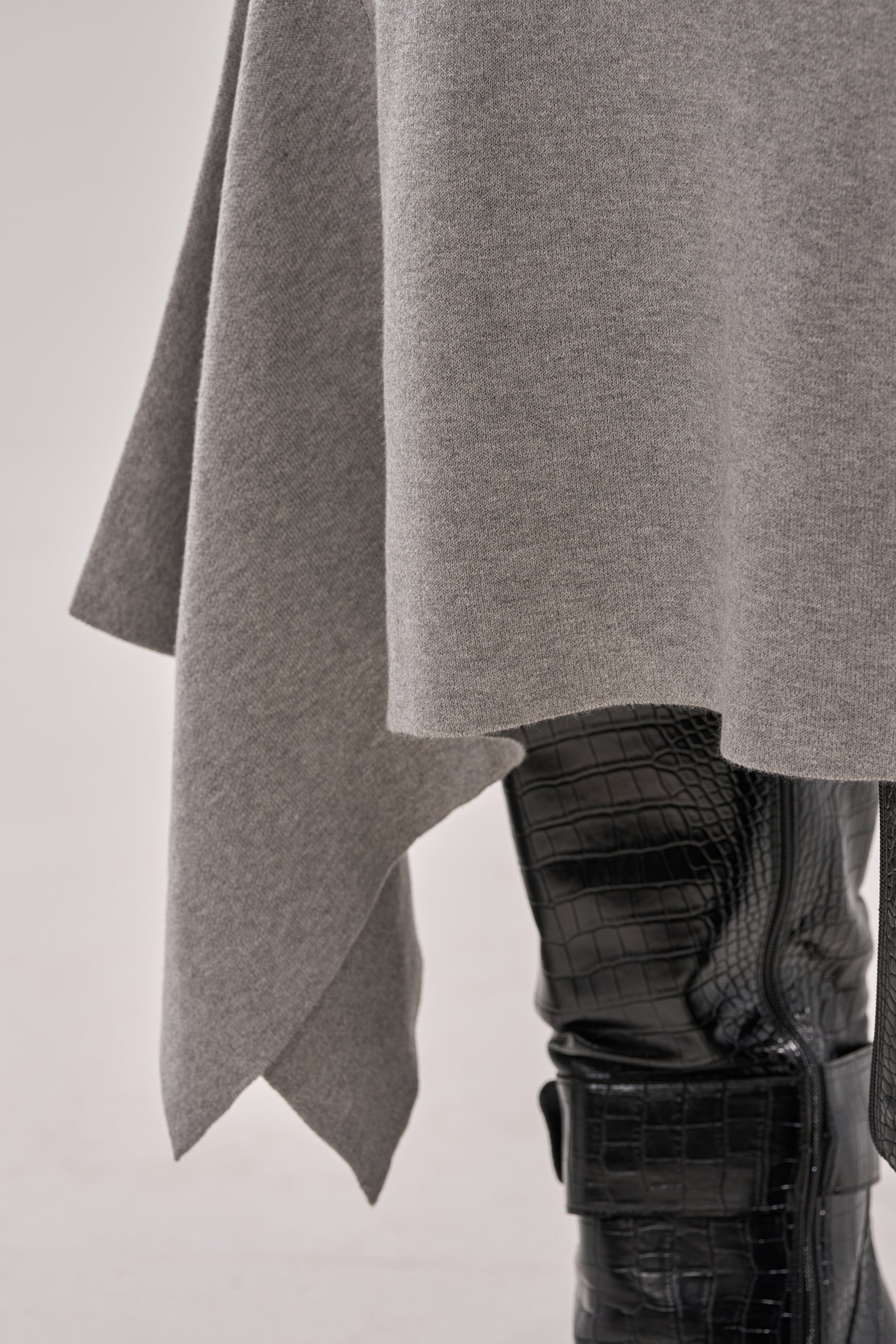Close-up of a person wearing an asymmetrical gray sweater with the LEXI MIDI SKIRT, featuring a high-rise fit and asymmetrical hem, contrasted by black crocodile-textured pants against a light background. 5