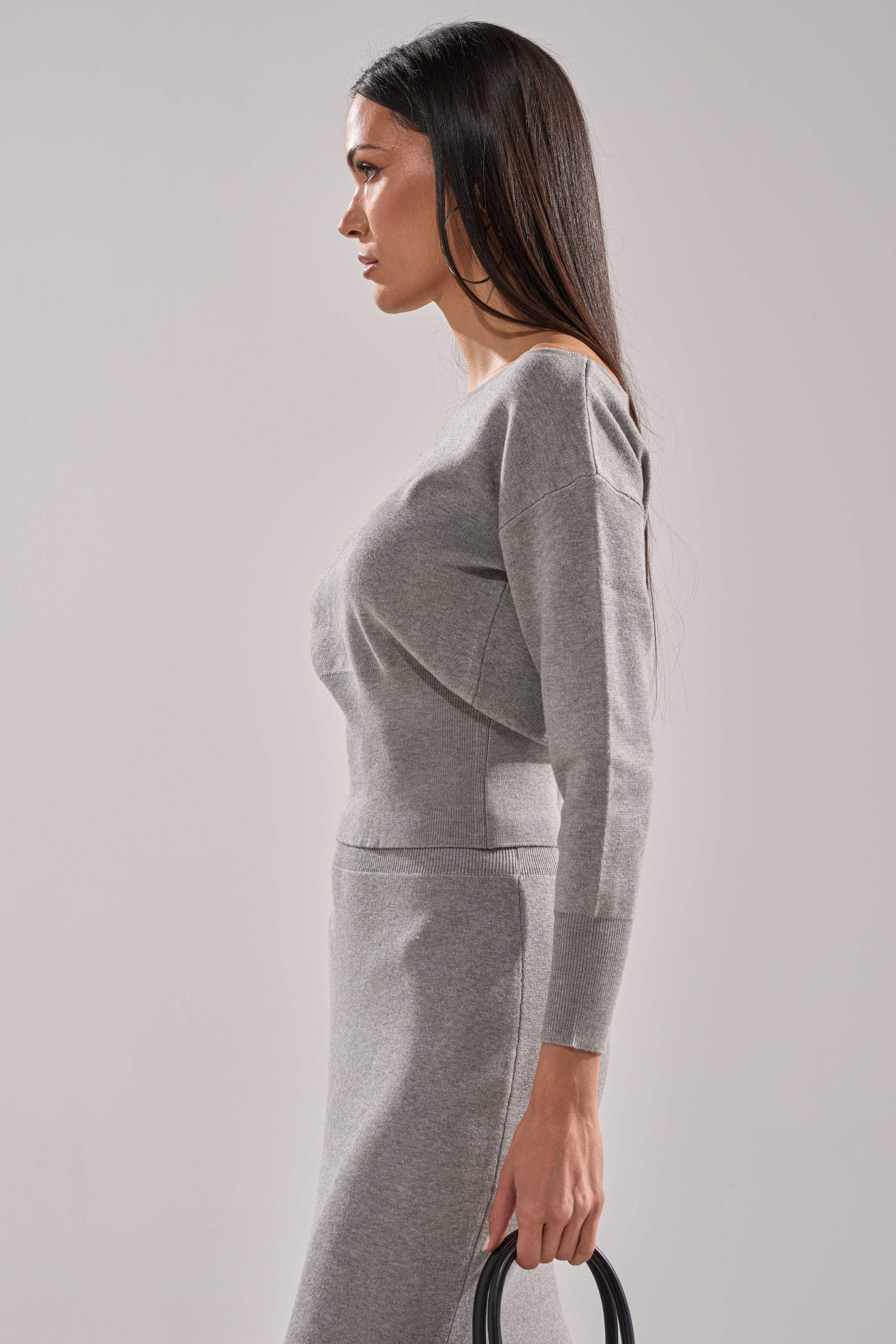 A woman with long dark hair stands in profile against a plain light gray background, wearing the AKIRA Label LEXI OFF THE SHOULDER SWEATER in light gray and holding a black circular handle bag. 3