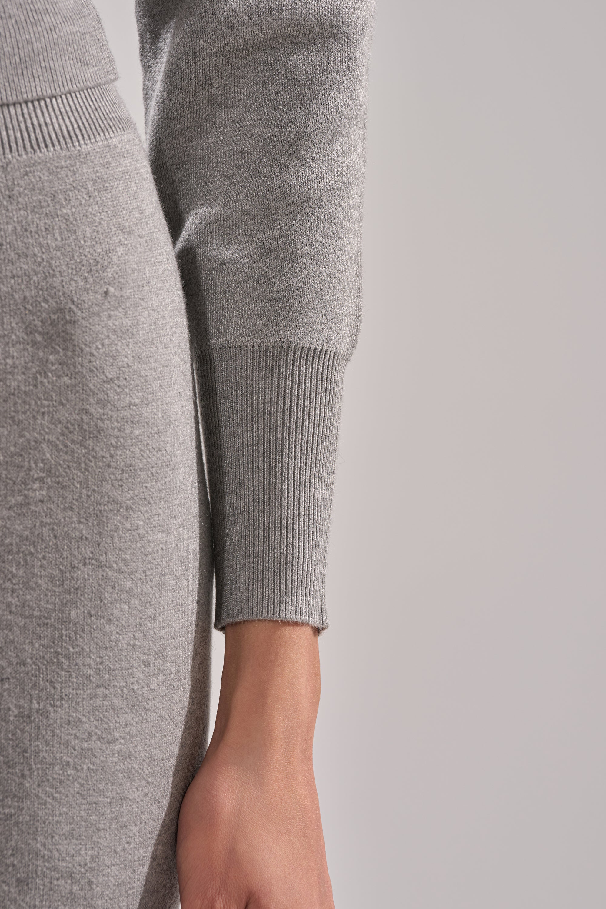 A close-up of a person's arm and hand wearing the LEXI OFF THE SHOULDER SWEATER in light gray stretch-knit with ribbed cuffs. The sweater is paired with matching pants against a plain, neutral background. 6