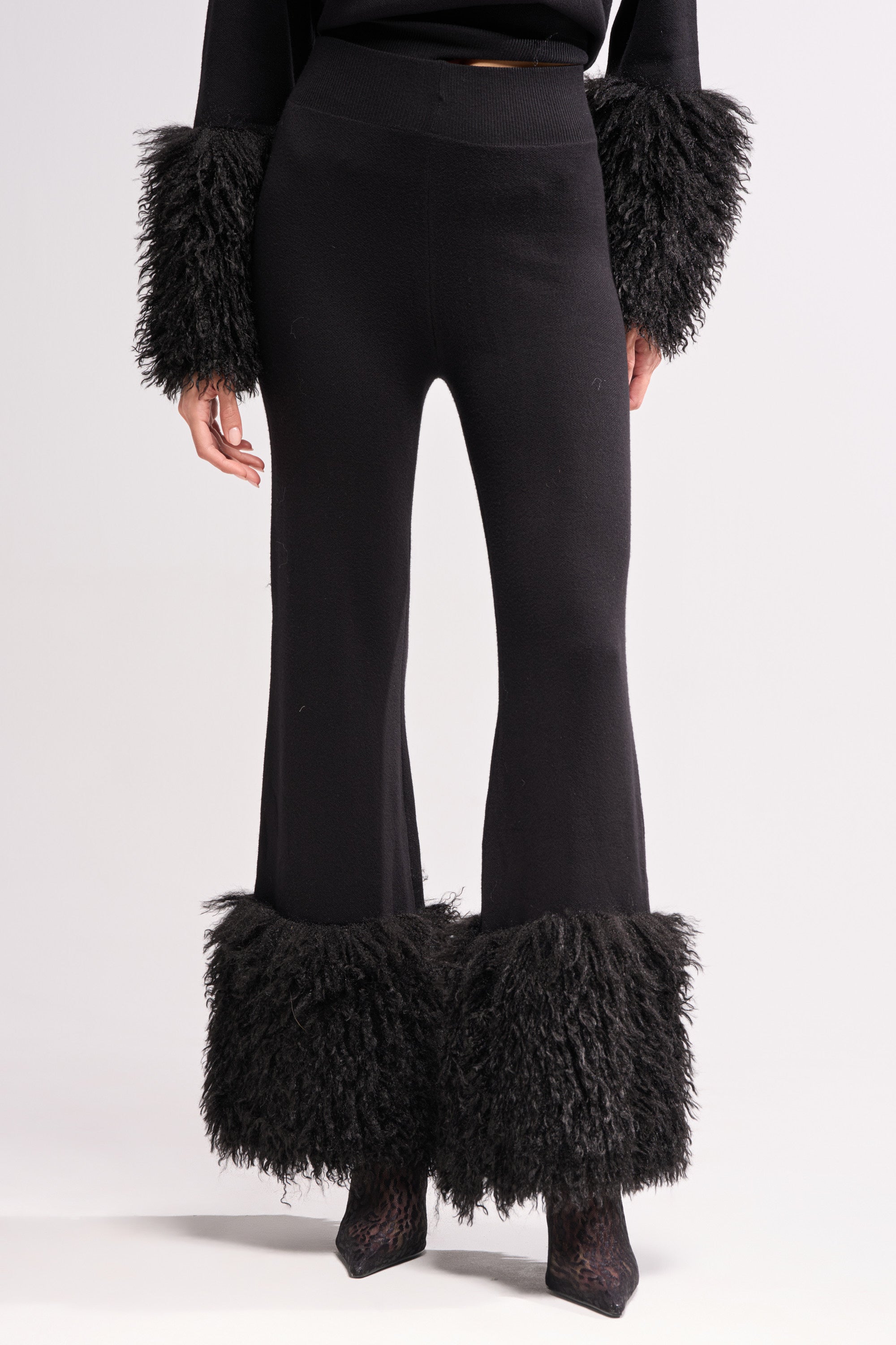 Someone wears the COZY VIBES SWEATER PANT WITH FUR CUFF IN BLACK—black fitted pants with wide-leg, textured faux fur cuffs—standing against a plain light background.