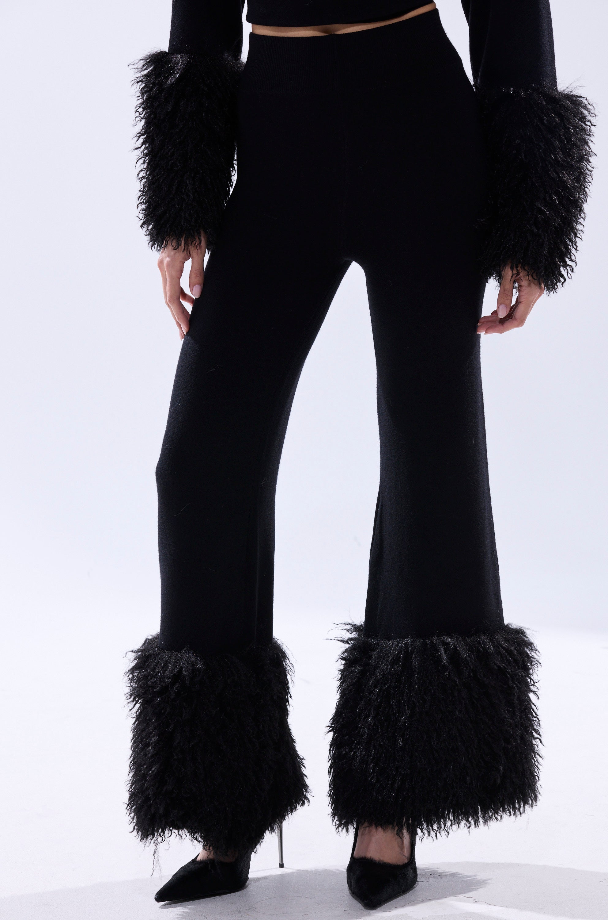 Wearing the COZY VIBES SWEATER PANT WITH FUR CUFF IN BLACK and a matching top with wide-leg design and furry black cuffs at wrists and ankles, paired with black pointed-toe heels, someone stands against a light background.