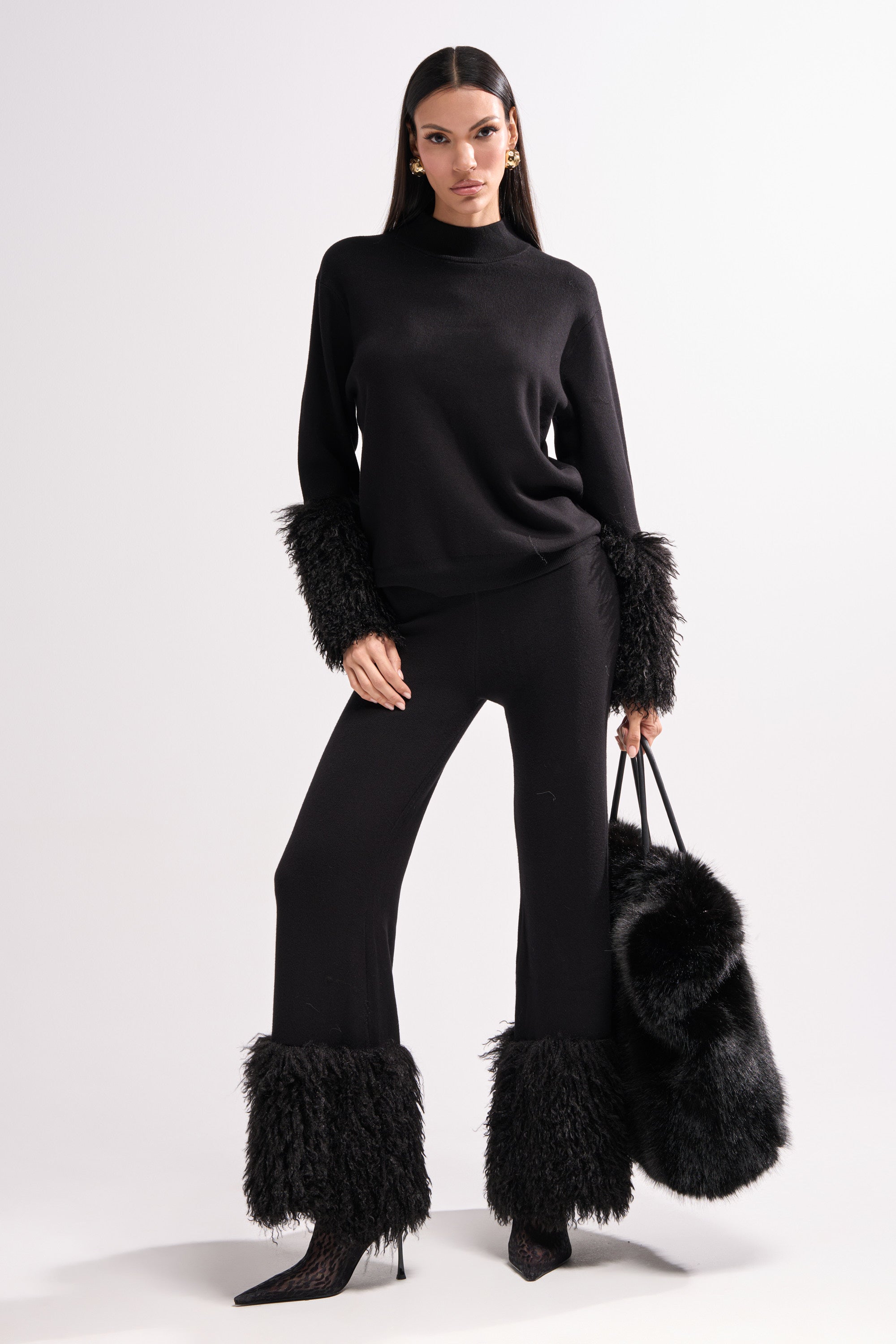 A woman stands confidently in the COZY VIBES SWEATER PANT WITH FUR CUFF IN BLACK, featuring wide legs and fur cuffs. She holds a large black fur bag, wears pointed-toe heels, and her long dark hair is straight against a plain white background.