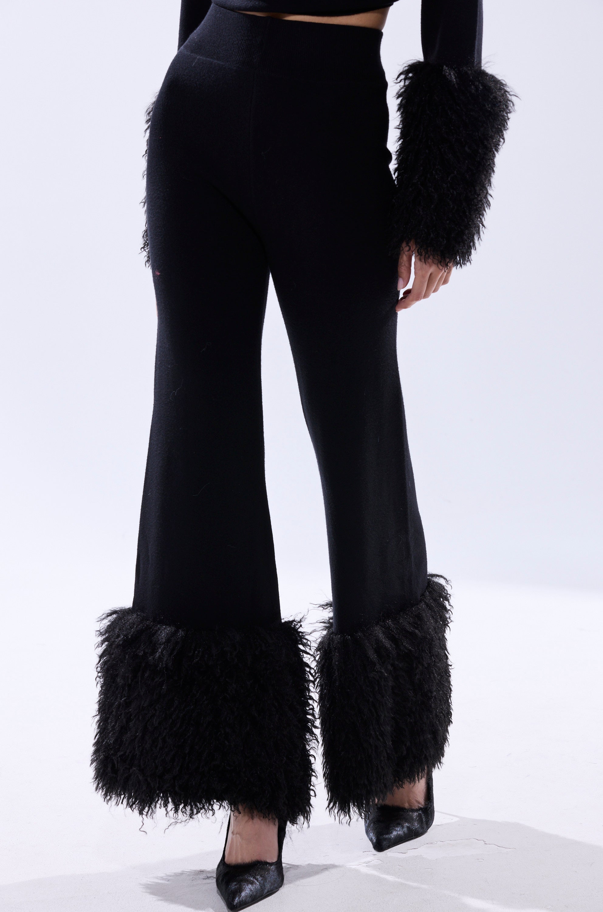 Wearing the COZY VIBES SWEATER PANT WITH FUR CUFF IN BLACK—featuring wide-leg black pants, shaggy fur cuffs, and matching fur-trimmed sleeves—a person poses against a light plain background in black pointed-toe heels.