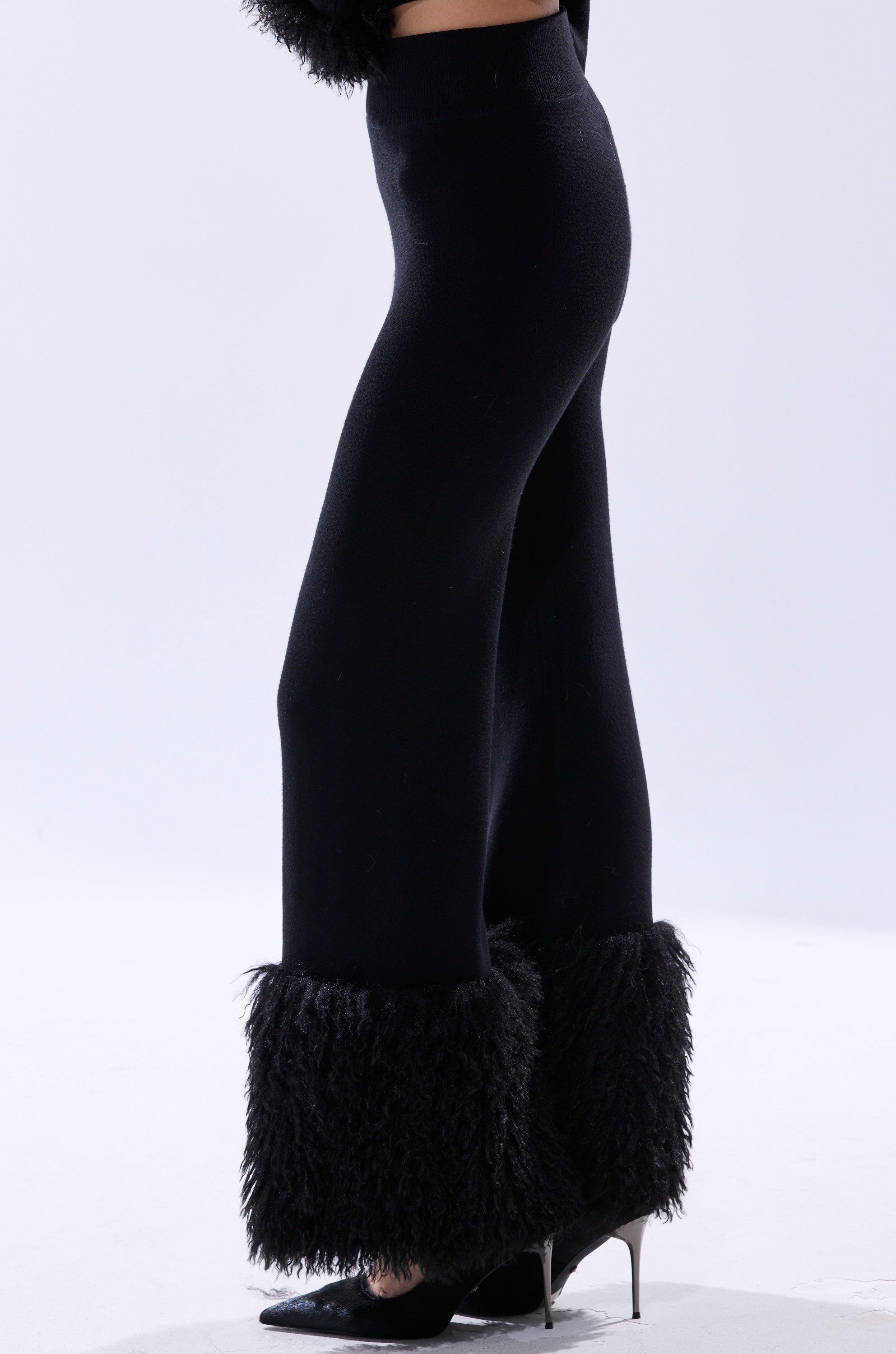A person in the COZY VIBES SWEATER PANT WITH FUR CUFF IN BLACK, featuring a wide-leg silhouette and fur cuffs, pairs it with black pointed-toe heels and stands against a plain light background.