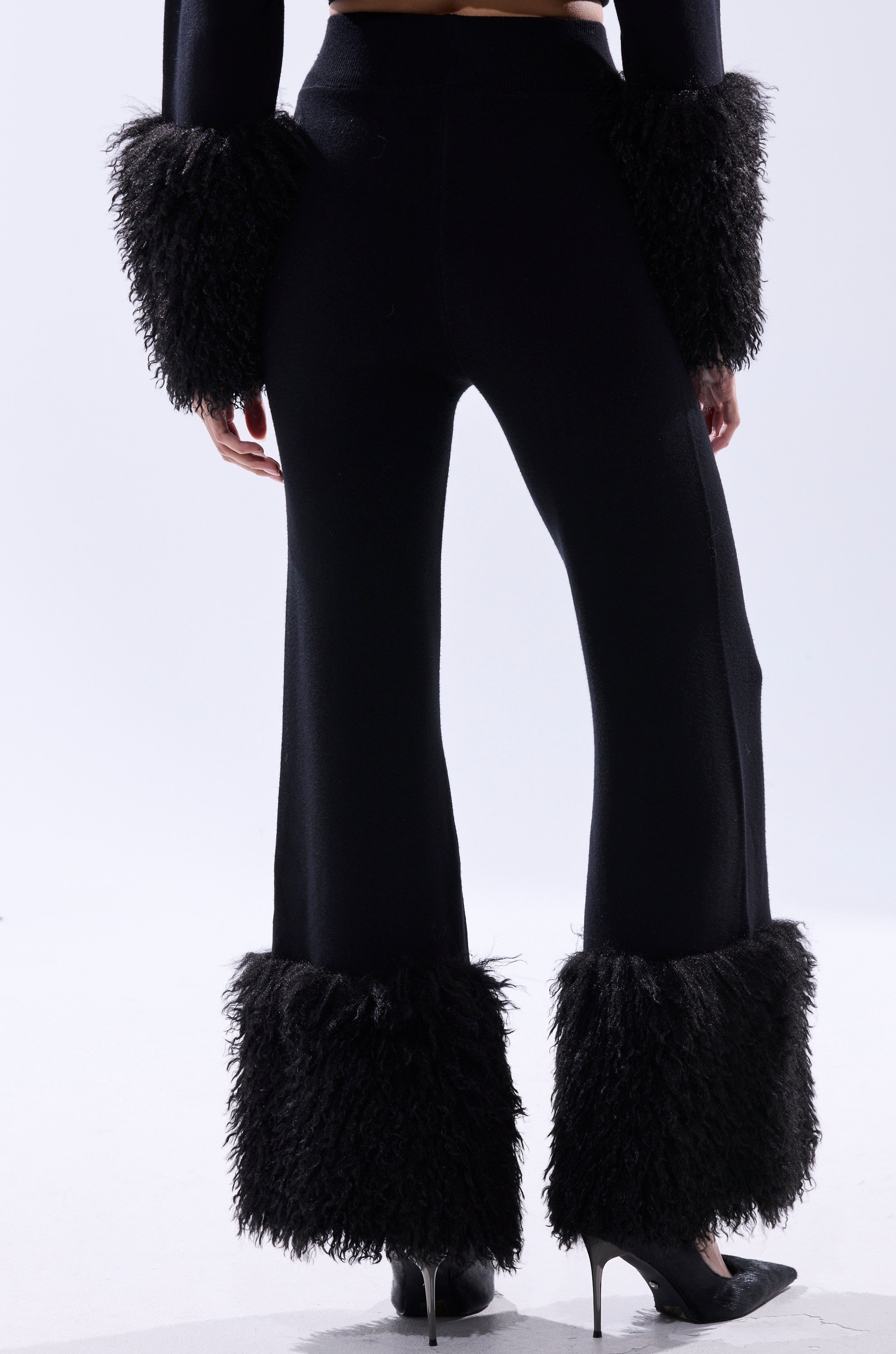 A person models the COZY VIBES SWEATER PANT WITH FUR CUFF IN BLACK—wide-leg black flared pants with bold fur cuffs—styled with pointed black heels, posed against a white background, channeling modern cozy vibes.