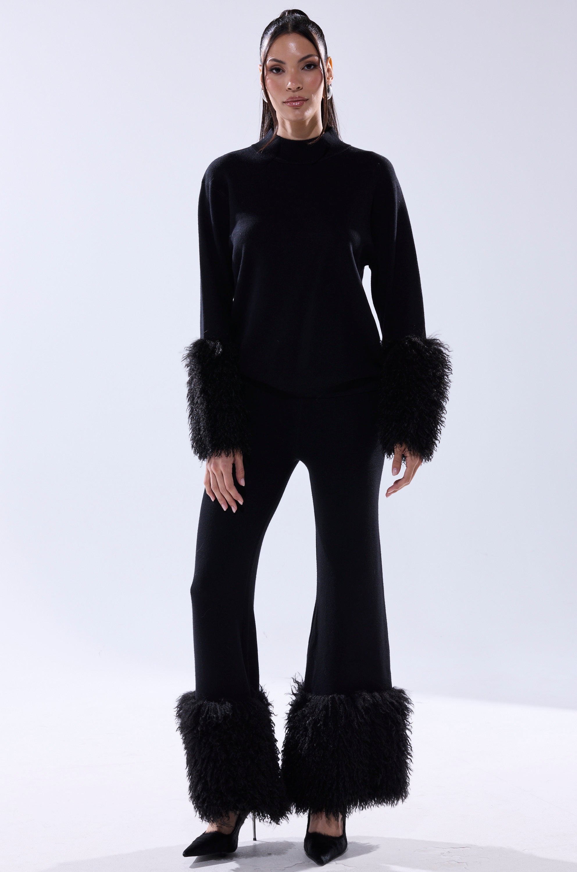 A woman wears the COZY VIBES SWEATER PANT WITH FUR CUFF IN BLACK—an outfit with wide-leg pants and long sleeves, both detailed with large, black fur cuffs. She pairs it with black heels and lets her long, straight dark hair down against a white background.