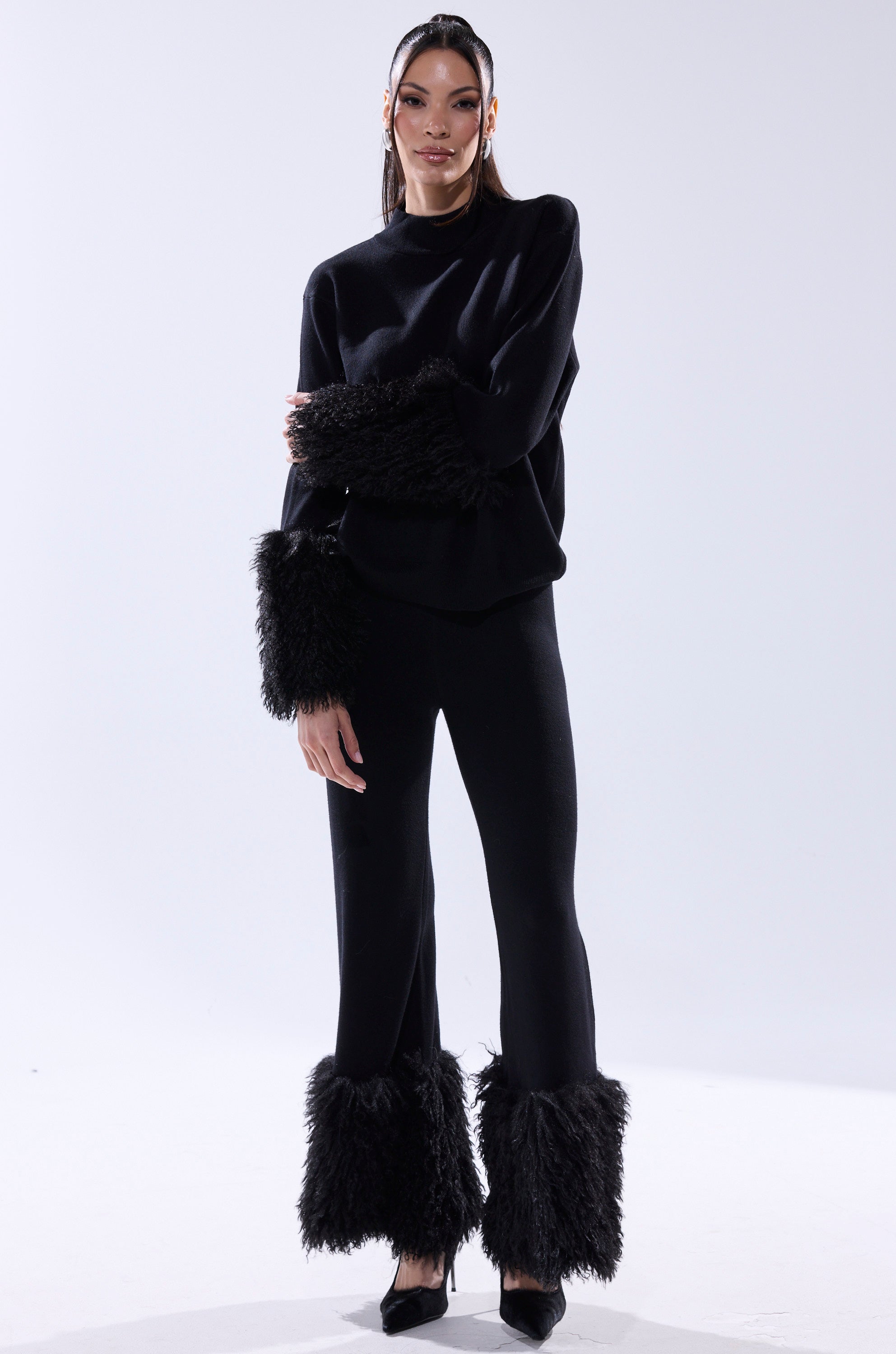 A woman with long hair and minimal jewelry poses confidently in the COZY VIBES SWEATER PANT WITH FUR CUFF IN BLACK—featuring wide legs and fur cuffs—paired with a matching black top and pointed-toe heels against a white background.