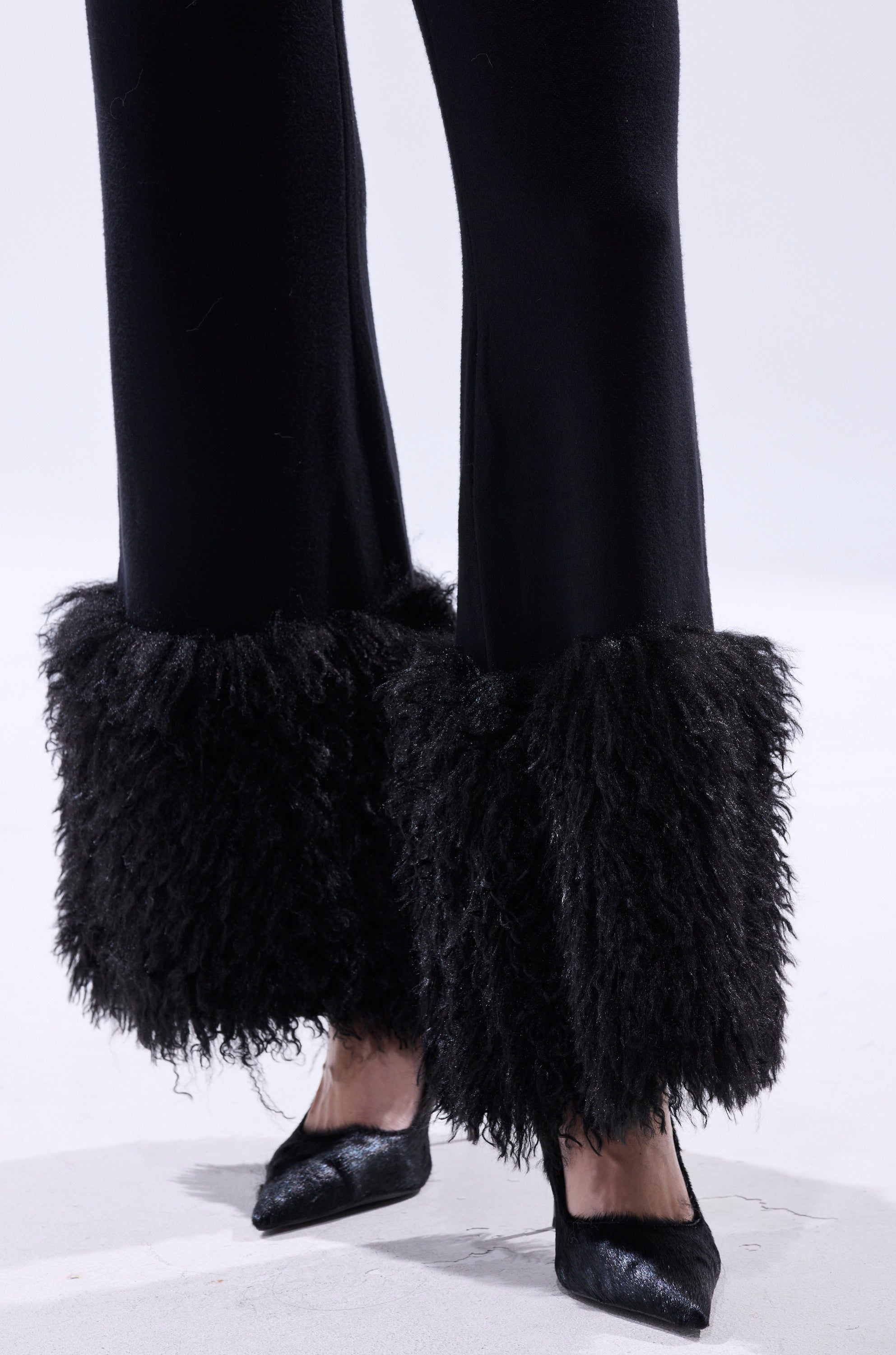 A person wears the COZY VIBES SWEATER PANT WITH FUR CUFF IN BLACK—wide-leg black pants with large, shaggy fur cuffs—paired with black pointed-toe heels, standing on a white surface.