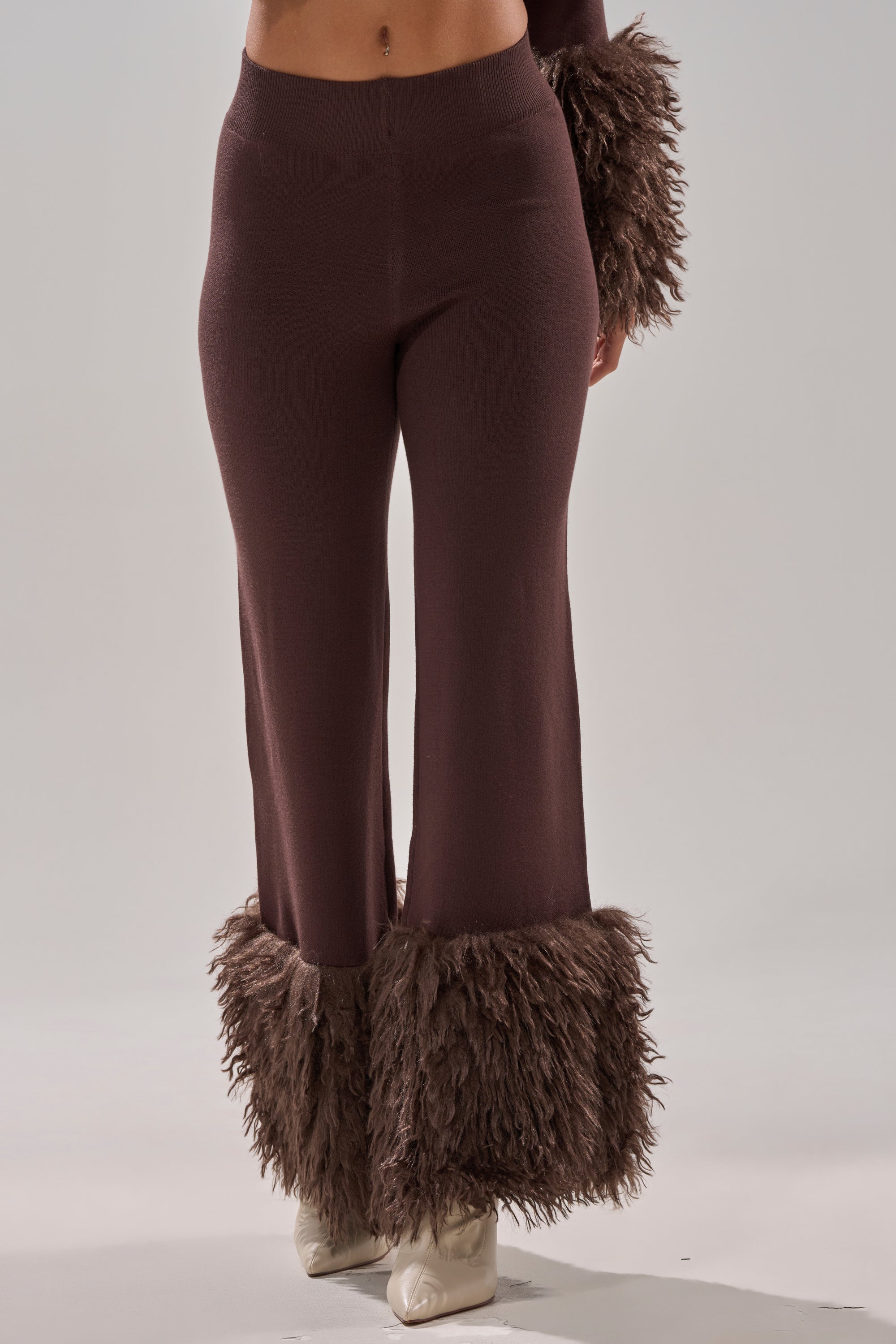 A person models the COZY VIBES SWEATER PANT WITH FUR CUFF IN BROWN, featuring wide legs and shaggy fur cuffs, shown from the waist down against a plain gray background. 2