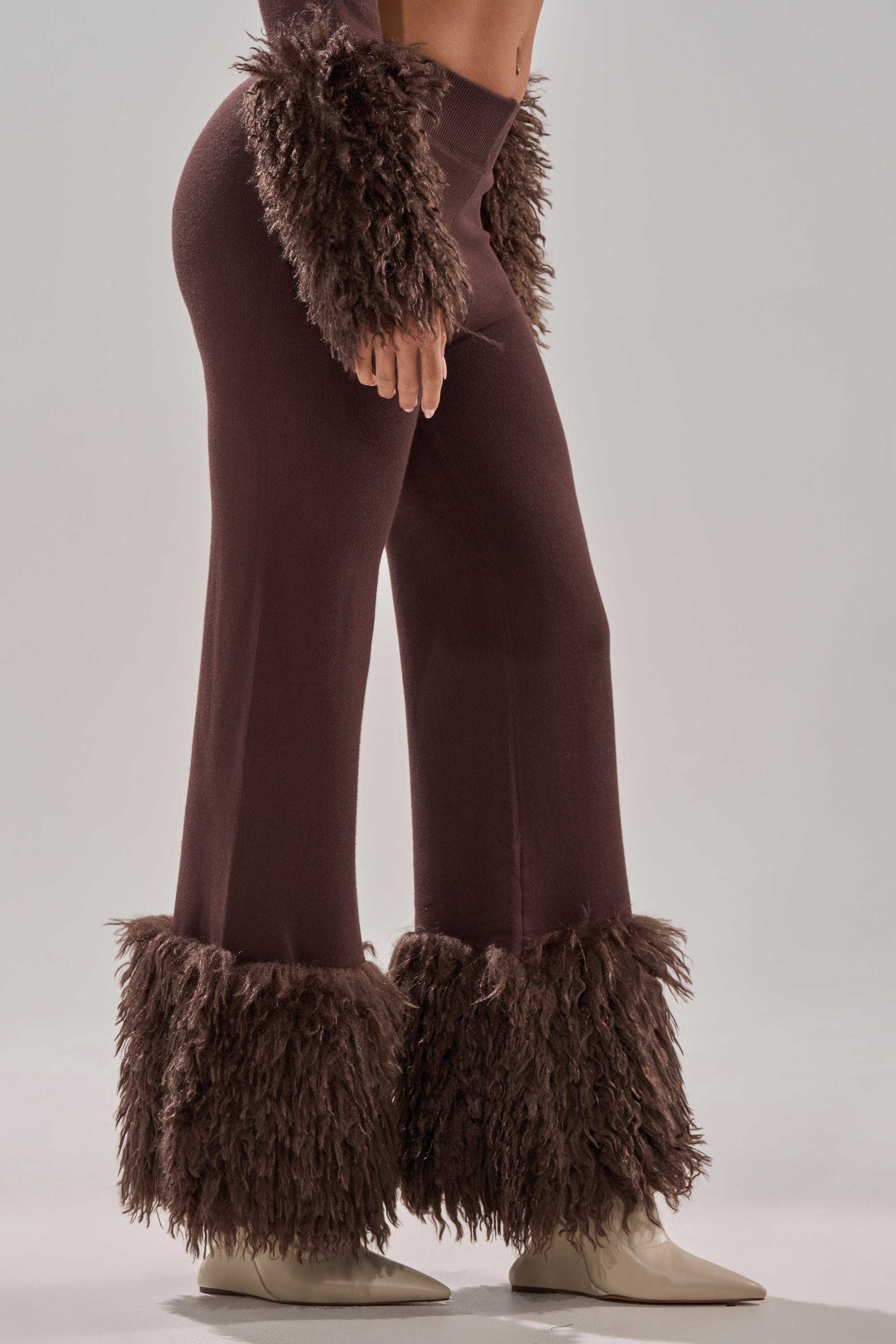 Wearing the COZY VIBES SWEATER PANT WITH FUR CUFF IN BROWN, featuring shaggy fur cuffs at the pant legs and sleeves, a person pairs it with light pointed-toe boots and stands against a plain gray background. 3