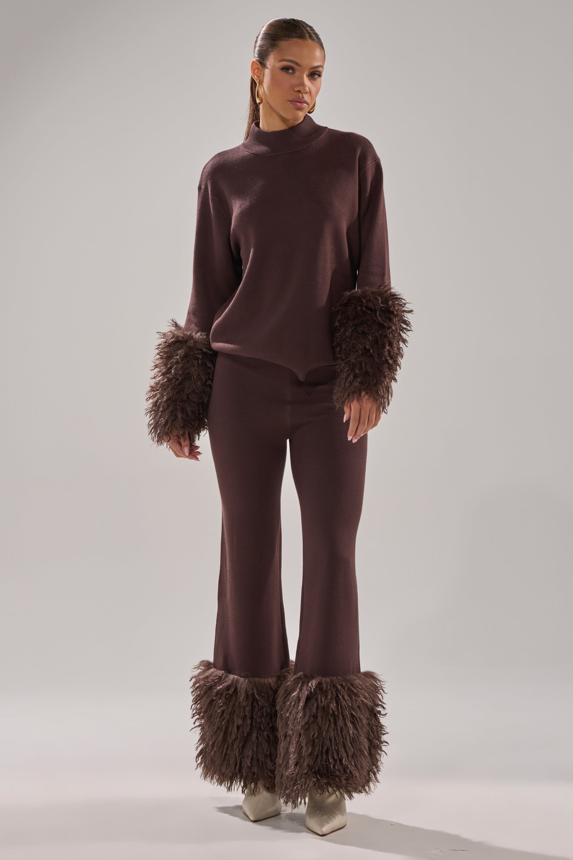 A woman models the COZY VIBES SWEATER PANT WITH FUR CUFF IN BROWN, featuring shaggy faux fur cuffs on the sleeves and pant hems, paired with white heeled boots against a plain light gray background. 5