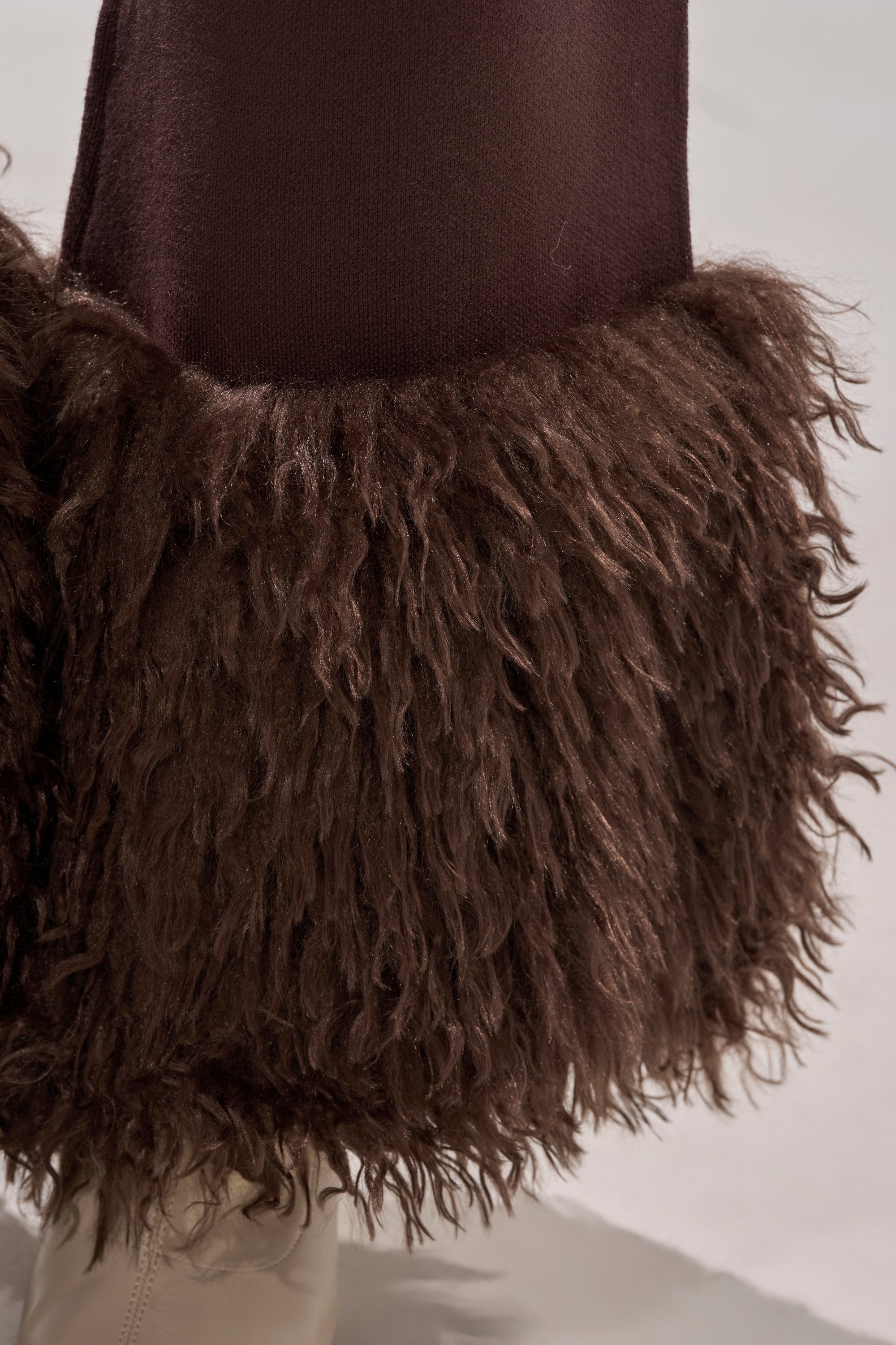 Close-up of the COZY VIBES SWEATER PANT WITH FUR CUFF IN BROWN, showing its textured, shaggy fur cuffs at the hem. A light shoe is visible below against a softly blurred neutral background. 6