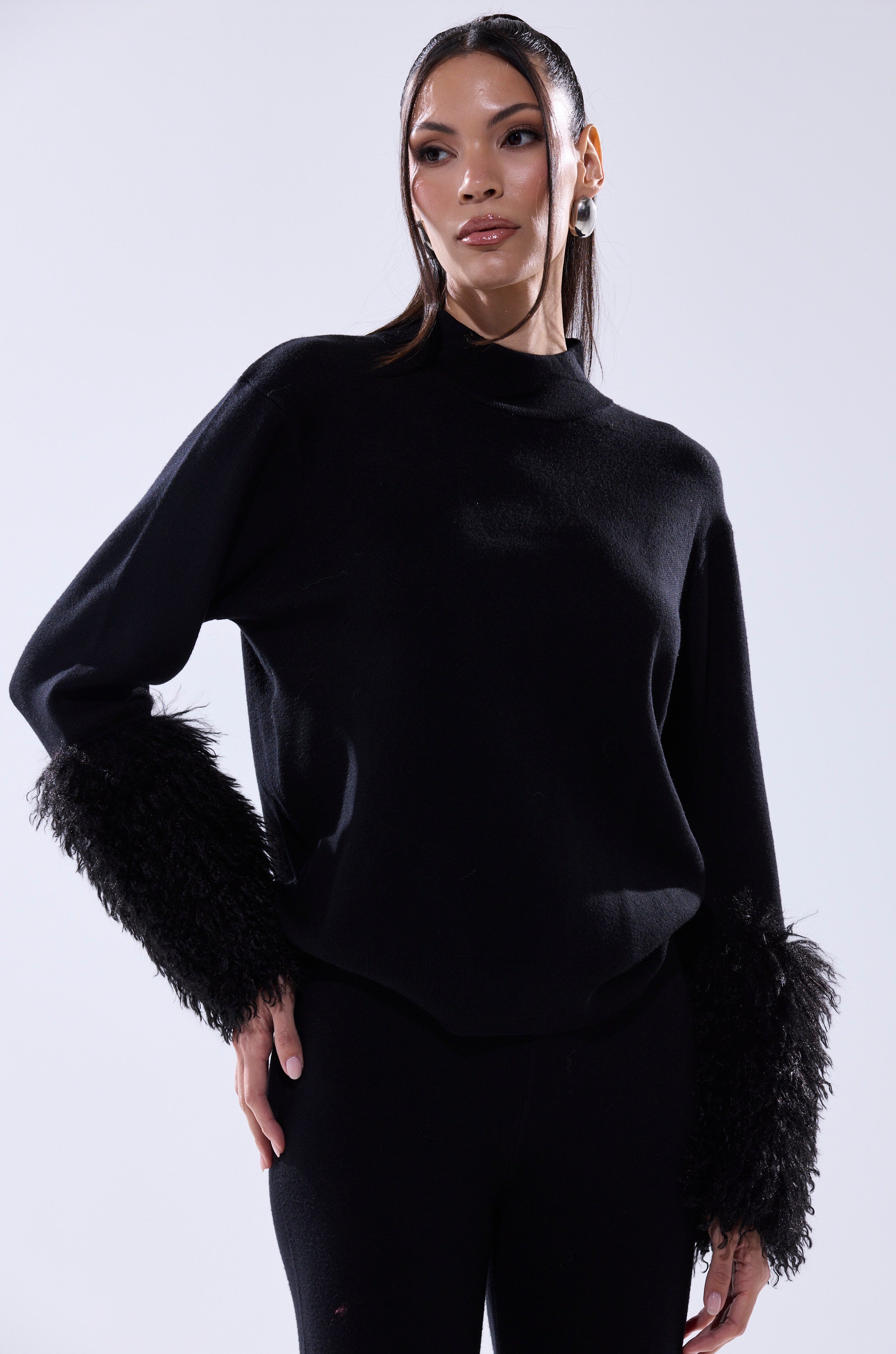 Woman models the COZY VIBES SWEATER WITH FUR CUFF IN BLACK, paired with black pants, standing against a light background with her hand on her hip.