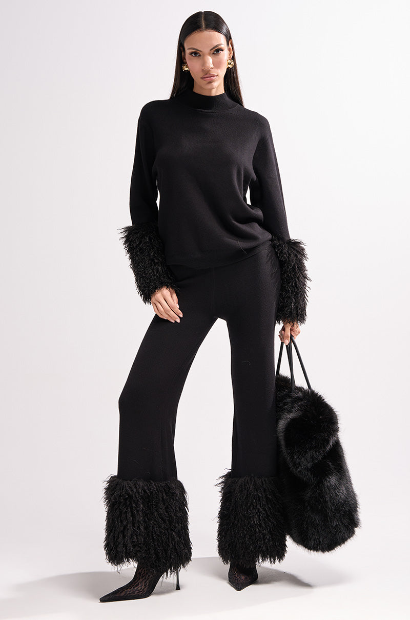 A woman poses confidently against a white background in the COZY VIBES SWEATER WITH FUR CUFF IN BLACK, paired with pants, heeled shoes, and a matching furry black bag, showcasing chic, cozy style.