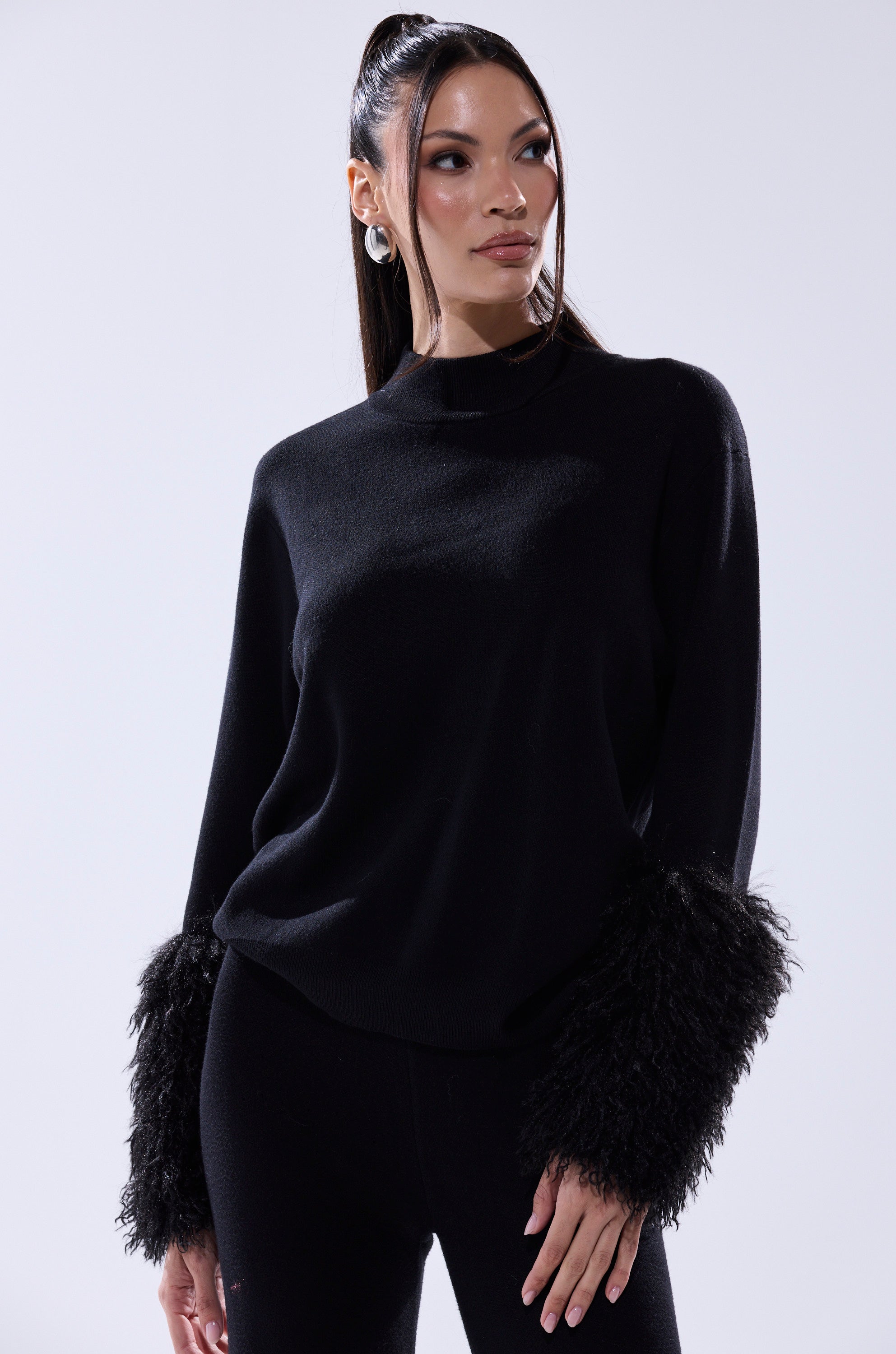 A woman in the COZY VIBES SWEATER WITH FUR CUFF IN BLACK and black pants stands against a plain light background, looking slightly to the side.