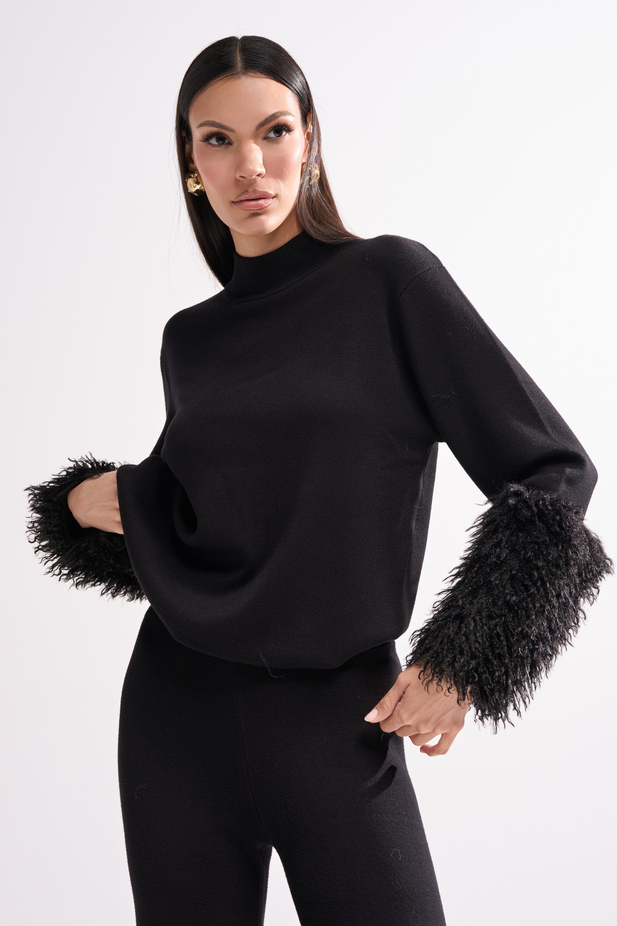 A woman with straight dark hair and gold earrings models the COZY VIBES SWEATER WITH FUR CUFF IN BLACK, featuring long sleeves with fur cuffs, against a plain white background.