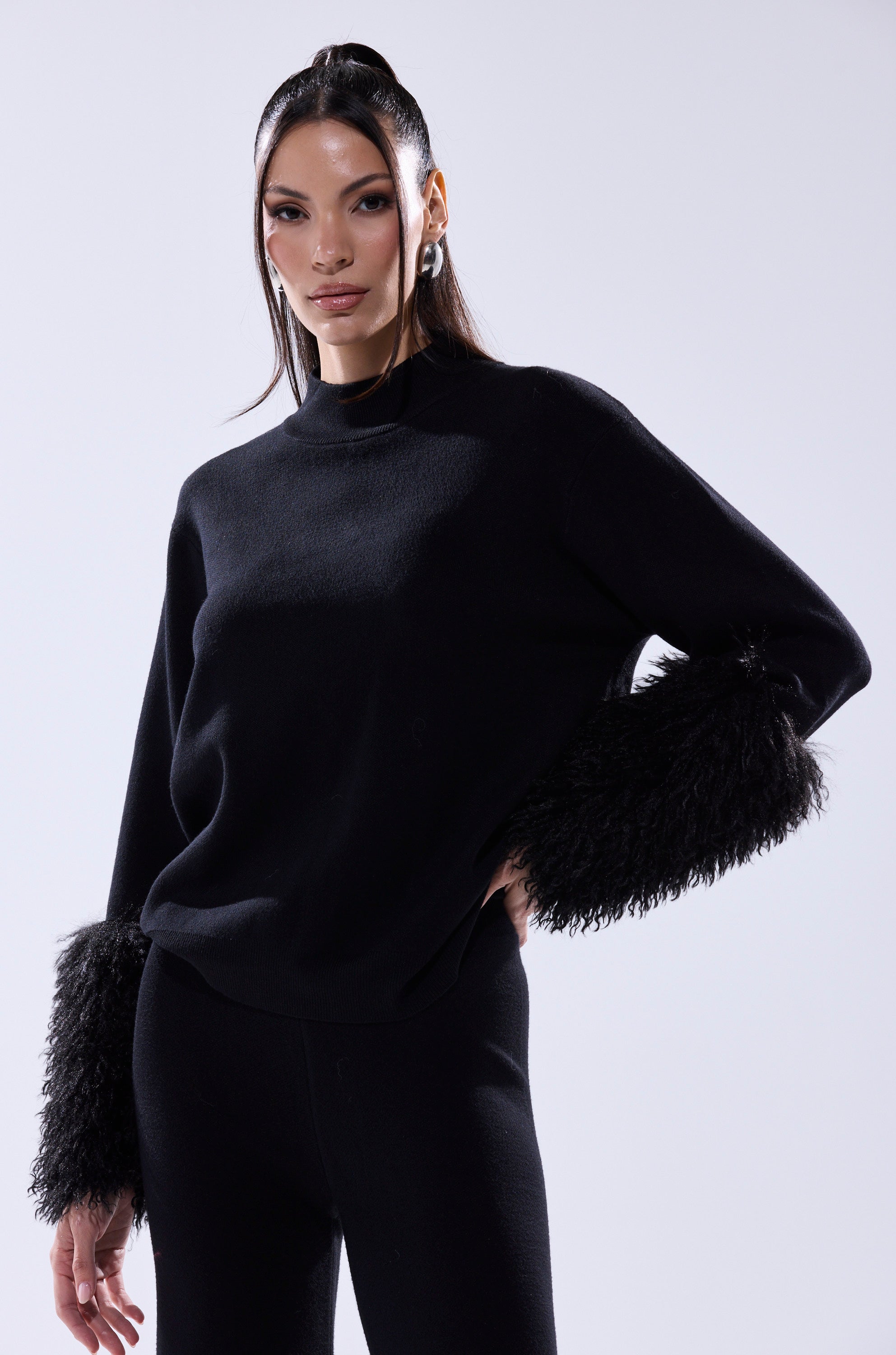 A woman models the COZY VIBES SWEATER WITH FUR CUFF IN BLACK—a pullover with fluffy fur cuffs—paired with matching black pants, standing confidently with one hand on her hip against a light background.
