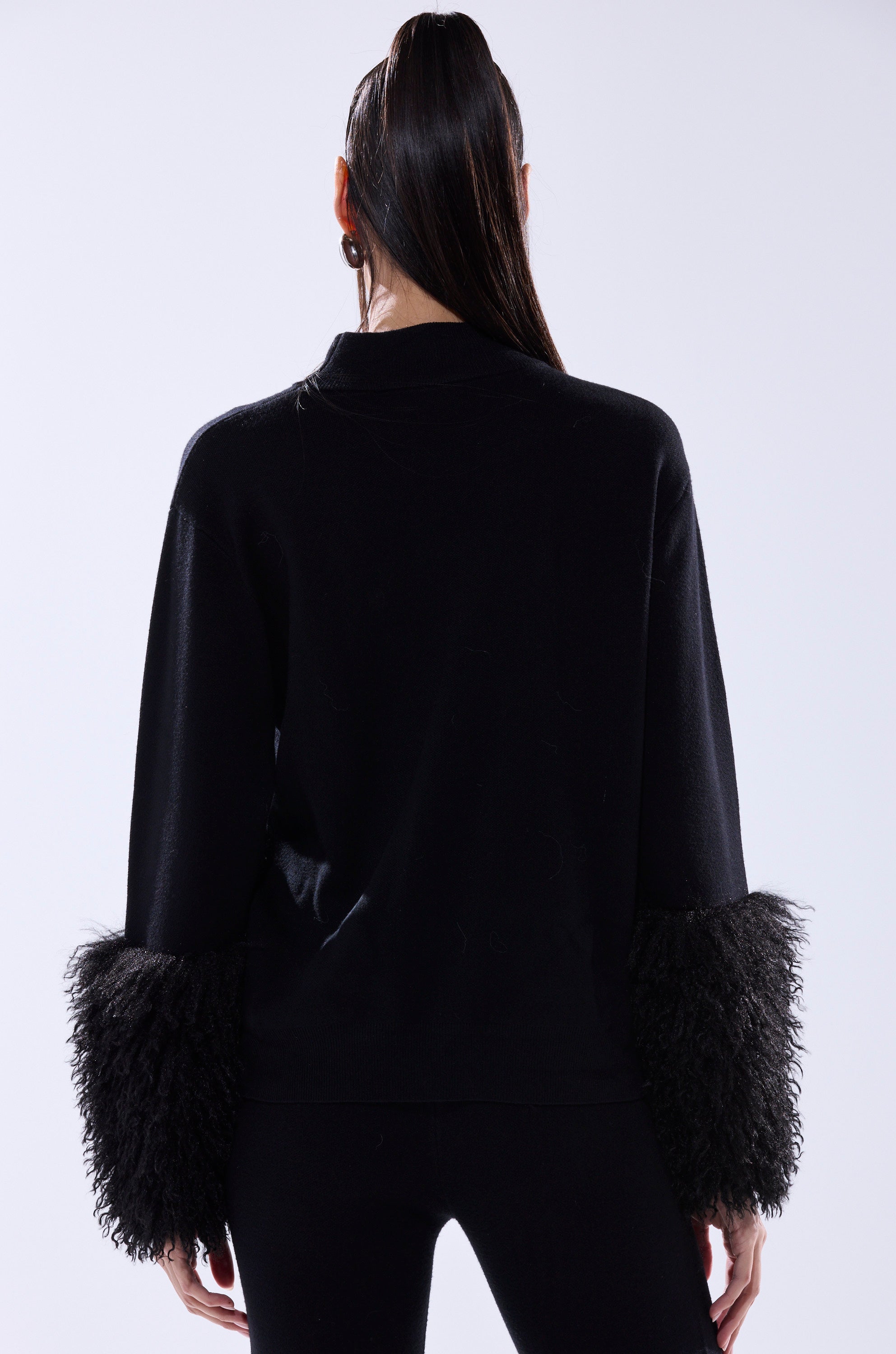 A person with long, dark hair in a high ponytail stands with their back to the camera, wearing the COZY VIBES SWEATER WITH FUR CUFF IN BLACK and matching black pants against a light background.