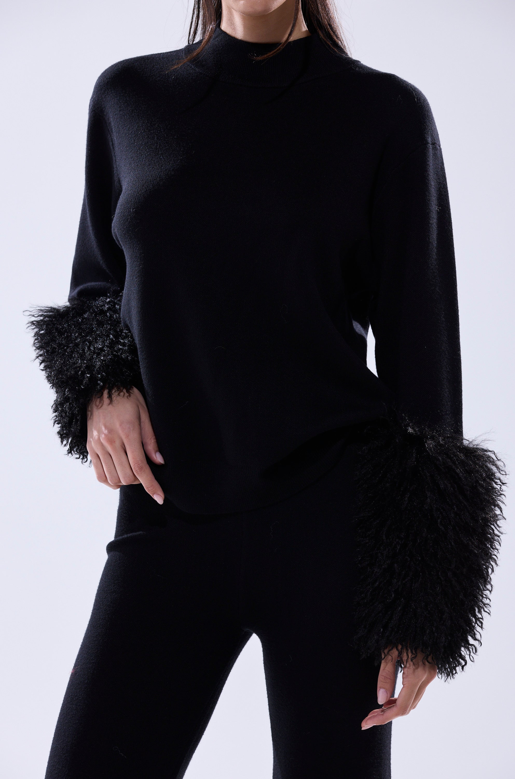 A person models the COZY VIBES SWEATER WITH FUR CUFF IN BLACK, a textured black pullover with fluffy fur cuffs, styled with matching black pants and posing with one hand on their hip against a light background.