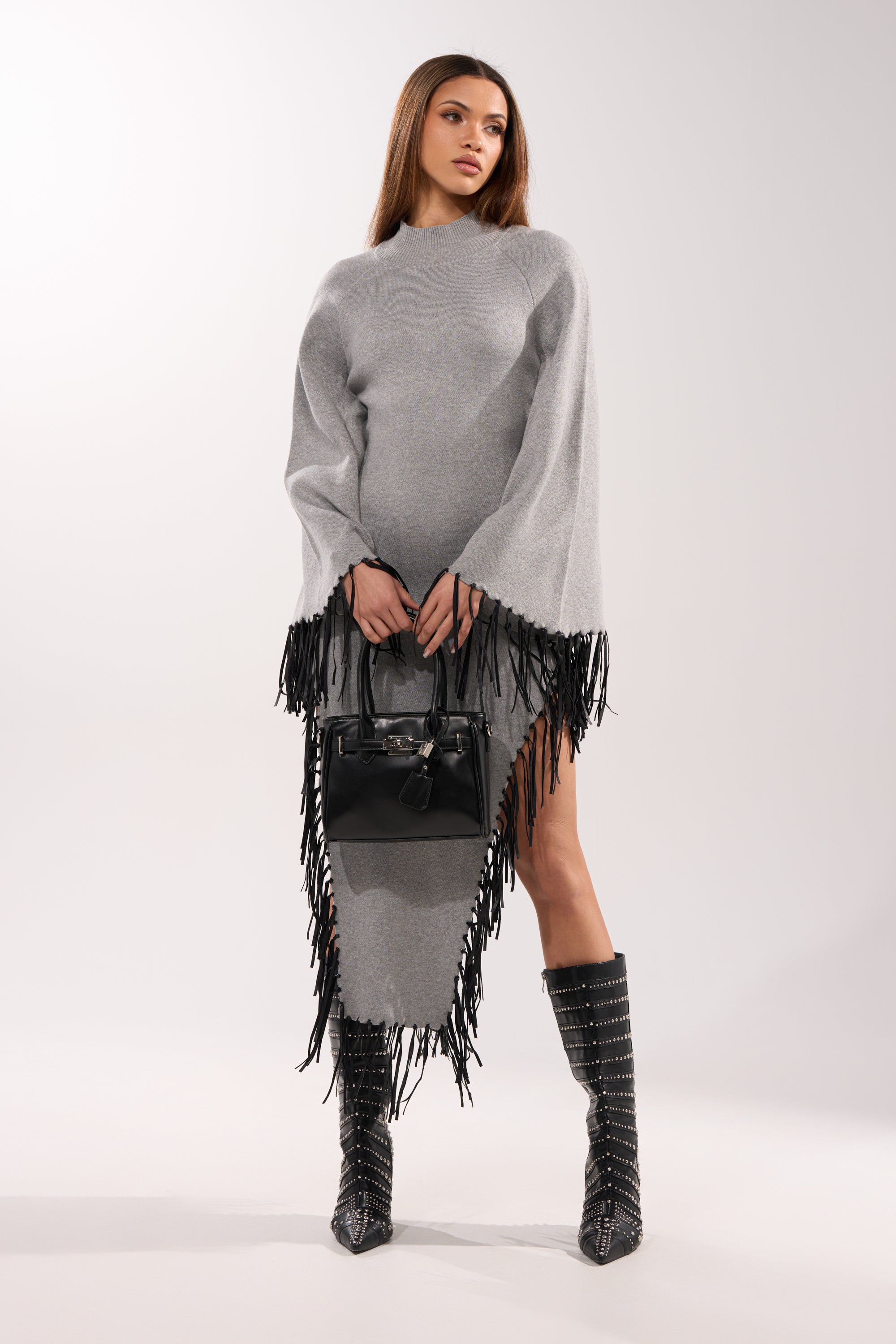 A woman wears the SWEATER WEATHER KNIT MIDI DRESS IN GREY, featuring subtle faux leather fringe, styled with studded black boots and a black handbag. The plain white background showcases her chic, modern look.