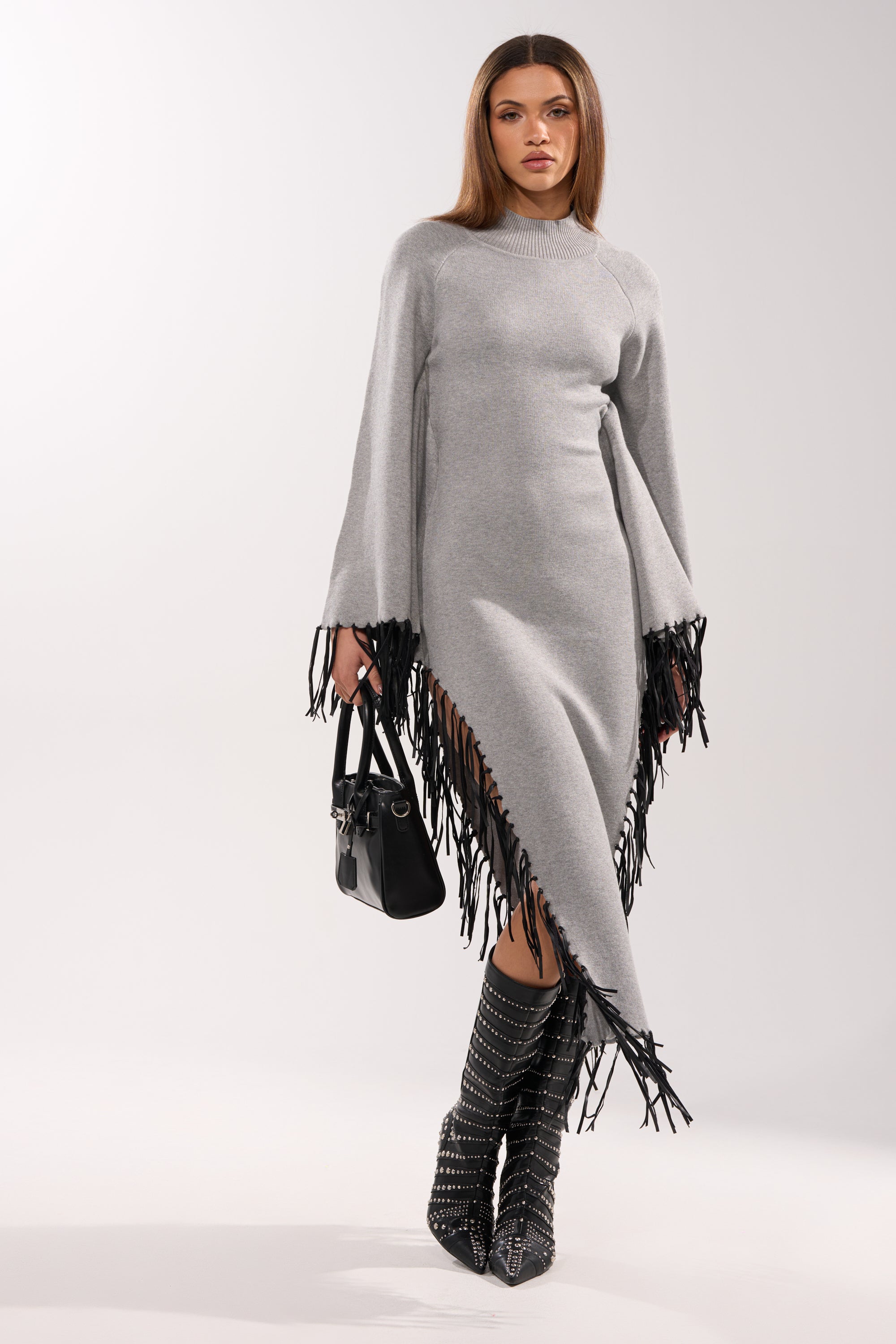 A woman wears the SWEATER WEATHER KNIT MIDI DRESS IN GREY, featuring an asymmetrical cut and black faux leather fringe on the hem and sleeves. She carries a black handbag, pairs it with studded knee-high black boots, against a plain white background.