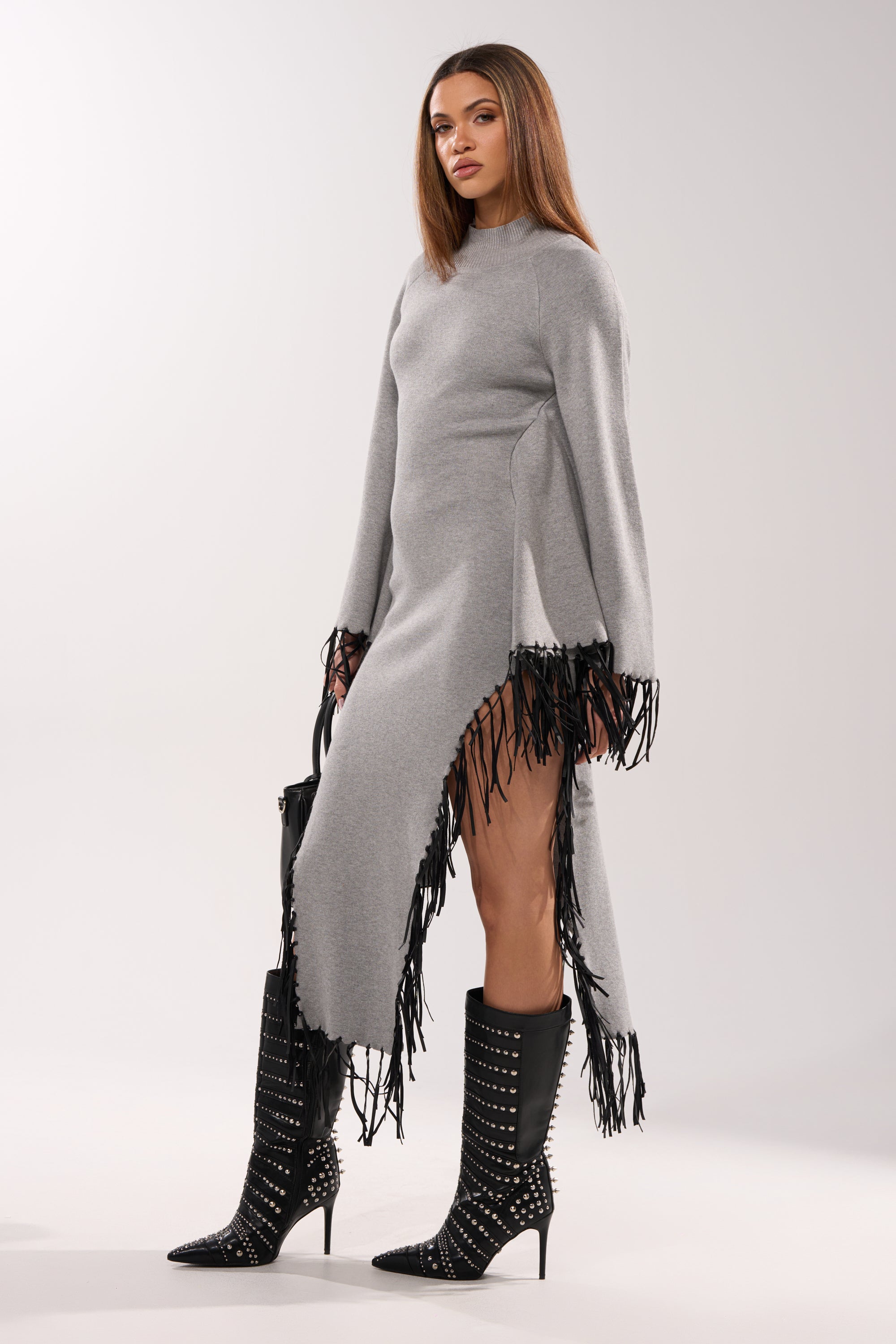 A woman stands confidently in the SWEATER WEATHER KNIT MIDI DRESS IN GREY, featuring faux leather fringe and high side slits. She styles it with black studded knee-high boots and a black bag against a light background.