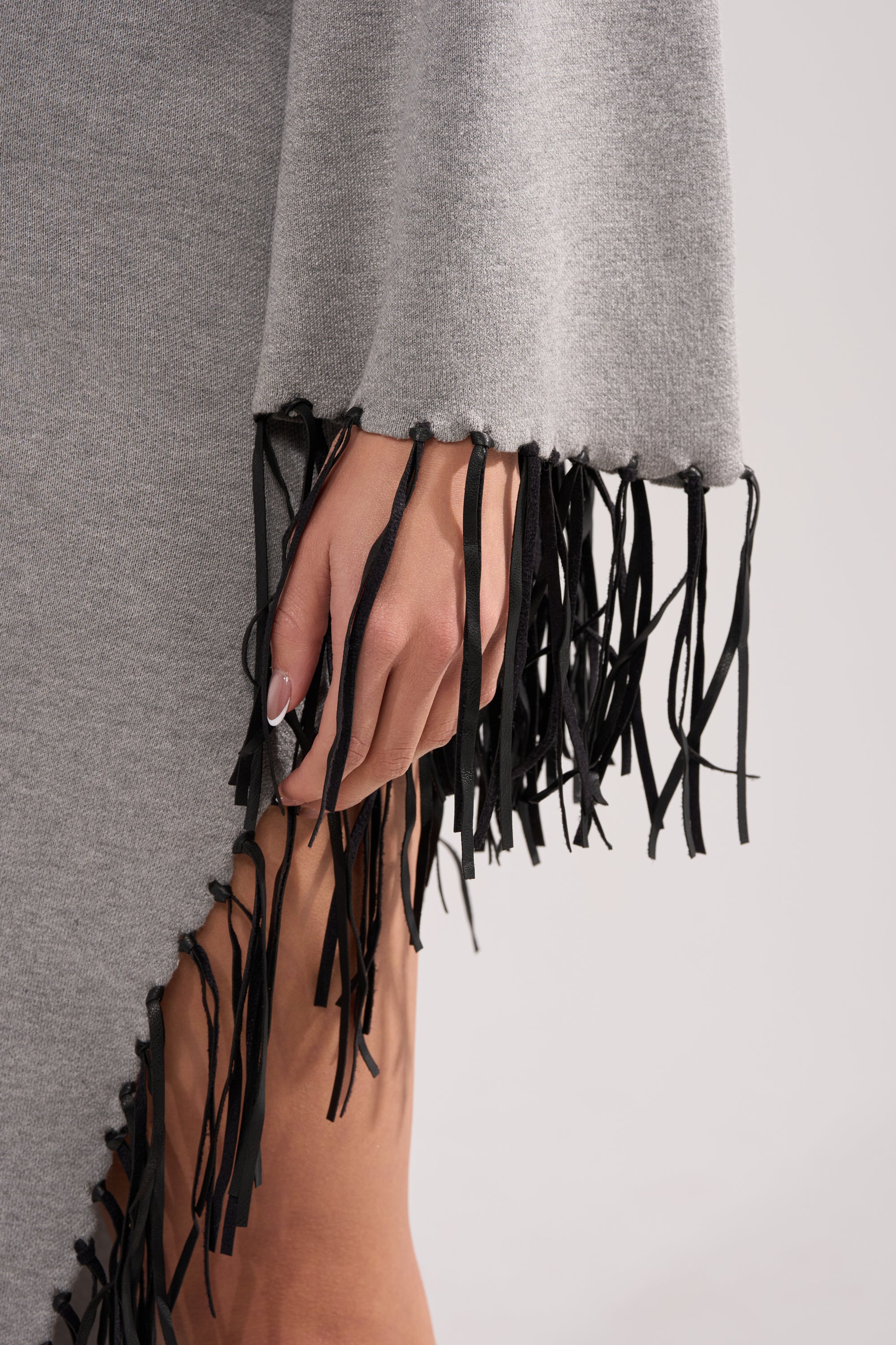 A close-up of a hand resting on the SWEATER WEATHER KNIT MIDI DRESS IN GREY, featuring black faux leather fringe along the sleeve and side, set against a plain background.