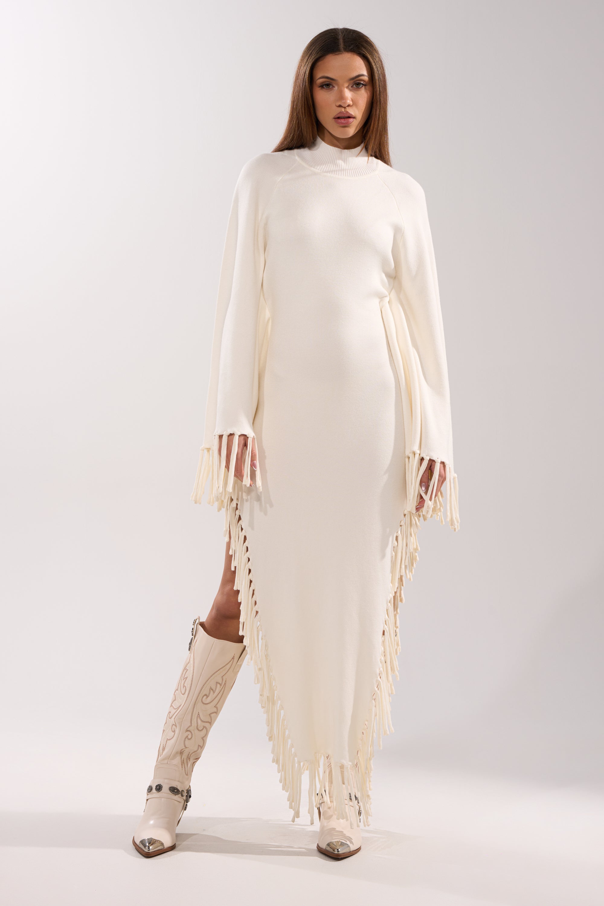 A woman wears the SWEATER WEATHER KNIT MIDI DRESS—a white knit midi with fringe trim and cape sleeves—styled with knee-high white cowboy boots. She has straight brown hair and a neutral expression against a plain backdrop.