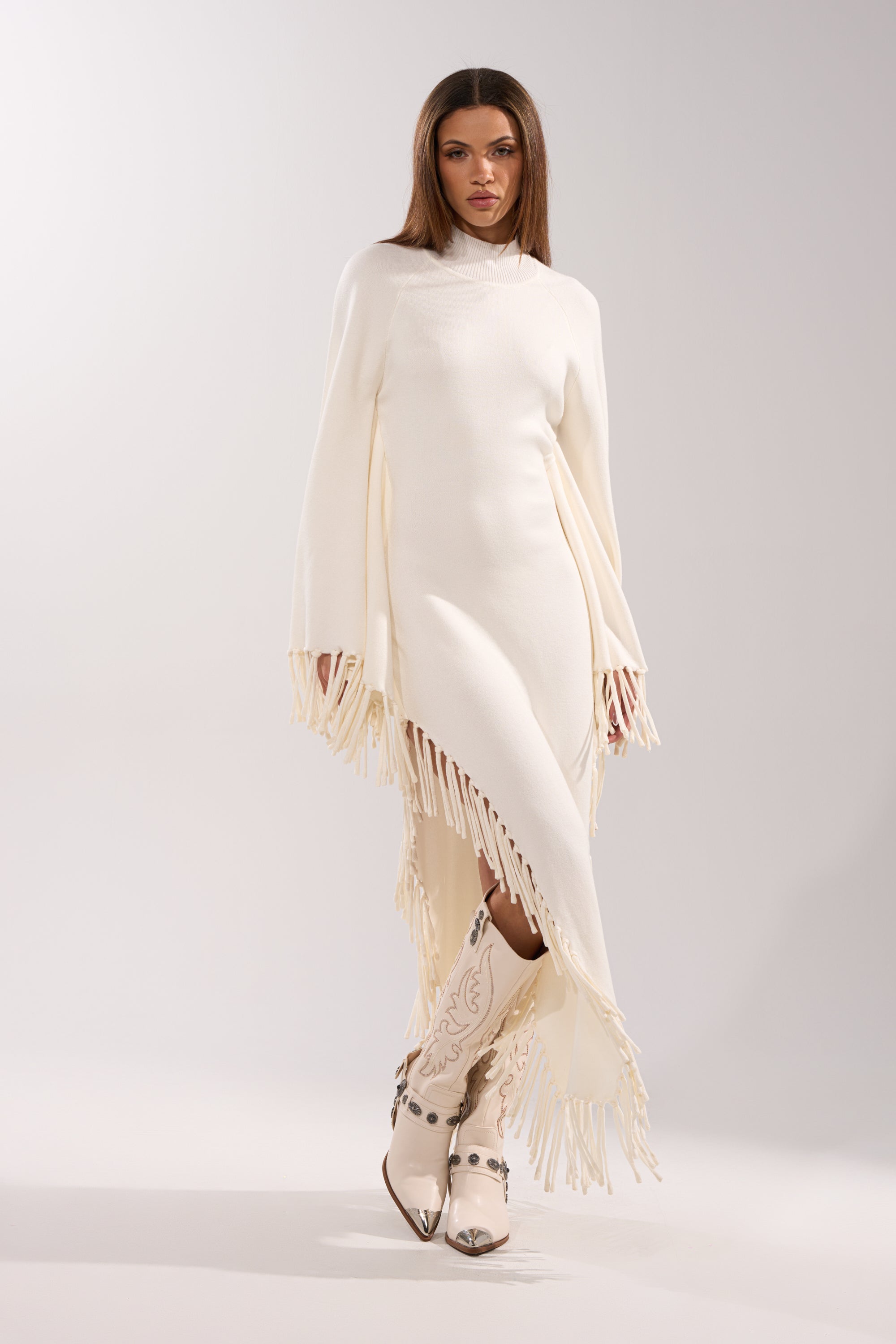 A woman poses confidently in the SWEATER WEATHER KNIT MIDI DRESS—an ivory fringe dress with cape sleeves—paired with white embroidered cowboy boots, set against a light gray background.