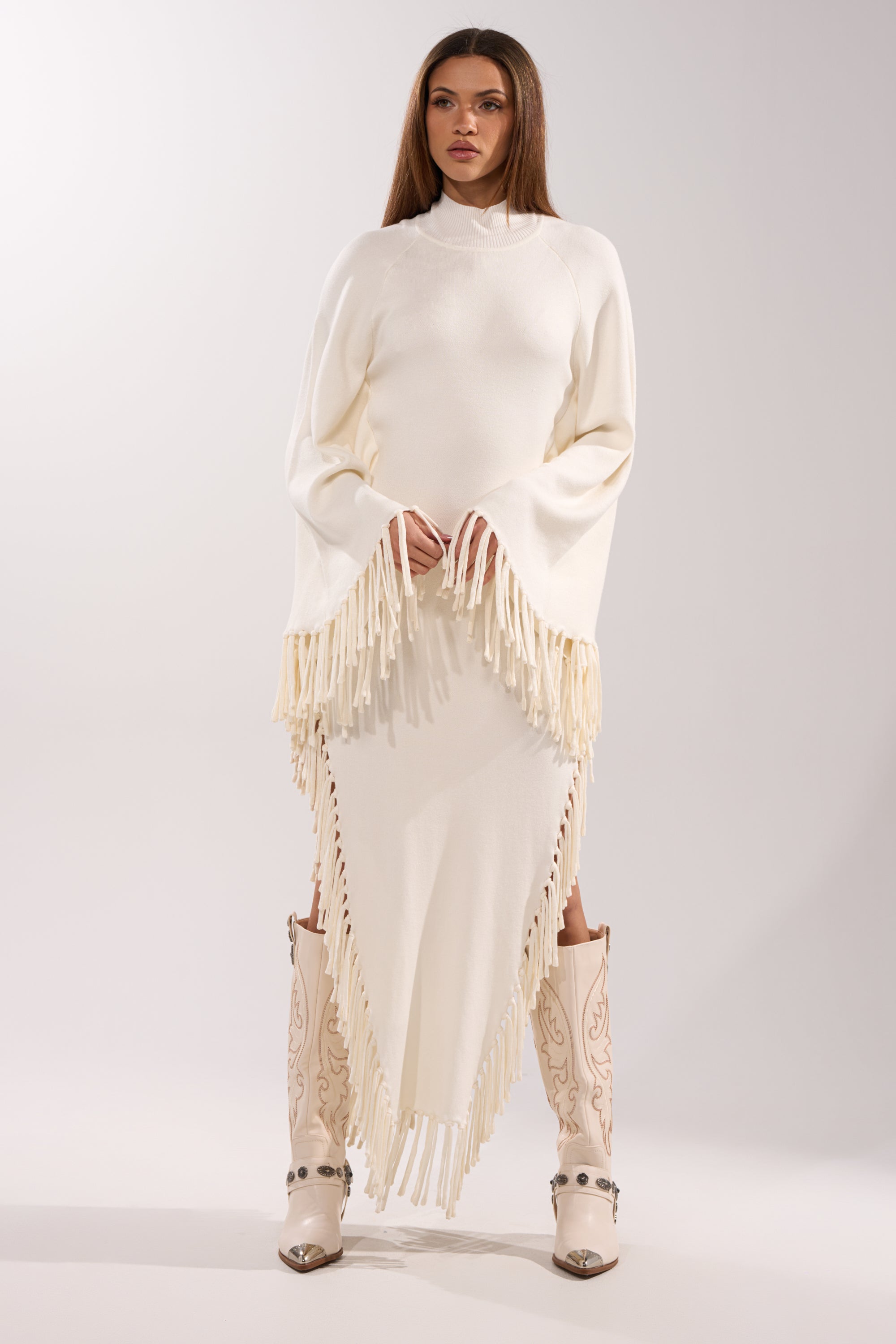 A woman faces forward in the SWEATER WEATHER KNIT MIDI DRESS, ivory with cape sleeves and decorative fringe. She pairs it with cream embroidered cowboy boots against a plain, light background.