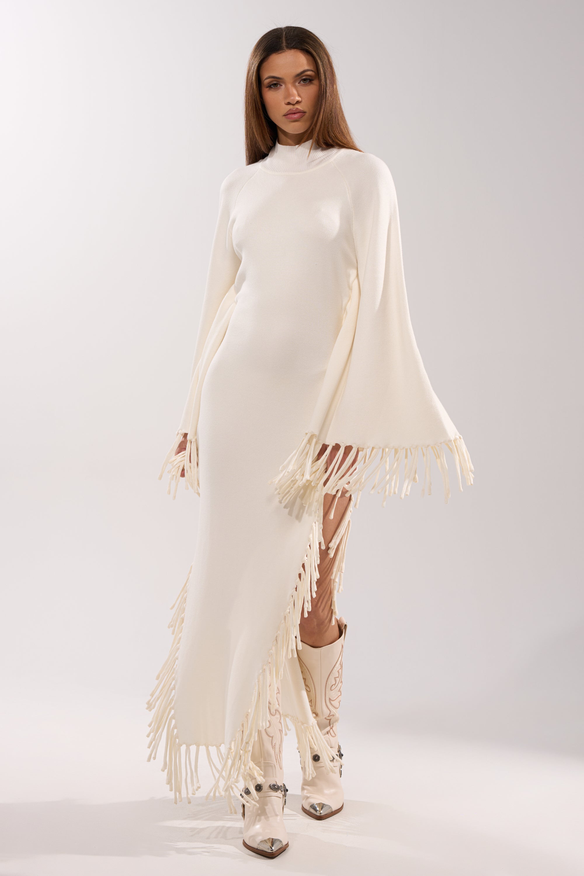 A woman poses confidently against a plain light background in the SWEATER WEATHER KNIT MIDI DRESS—a fitted white midi with cape sleeves and fringe trim—paired with white cowboy boots featuring decorative stitching.