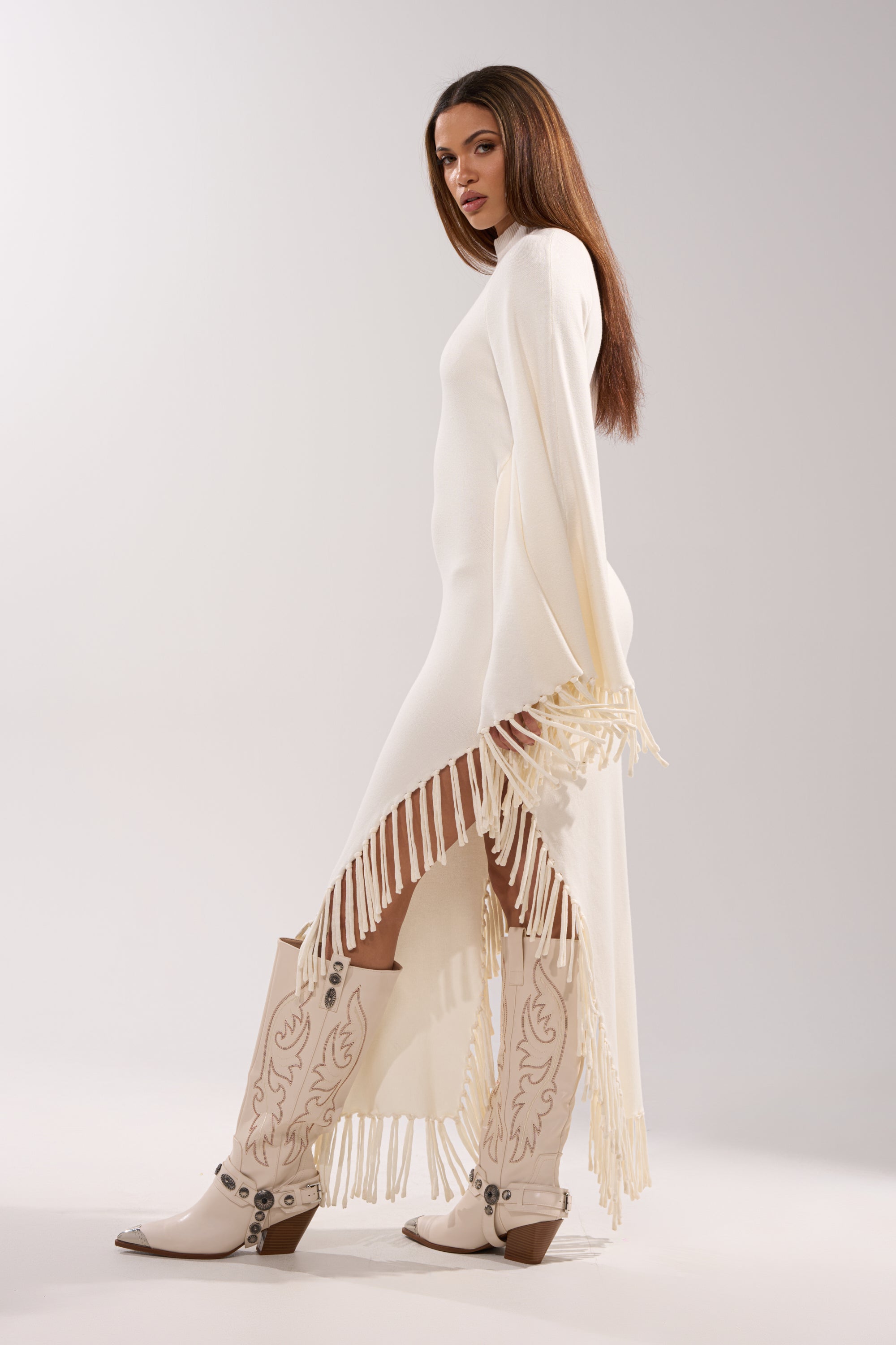 A woman poses sideways in the SWEATER WEATHER KNIT MIDI DRESS, a fitted white midi with side fringe trim. She pairs it with white cowboy boots featuring decorative stitching, standing against a plain light background.