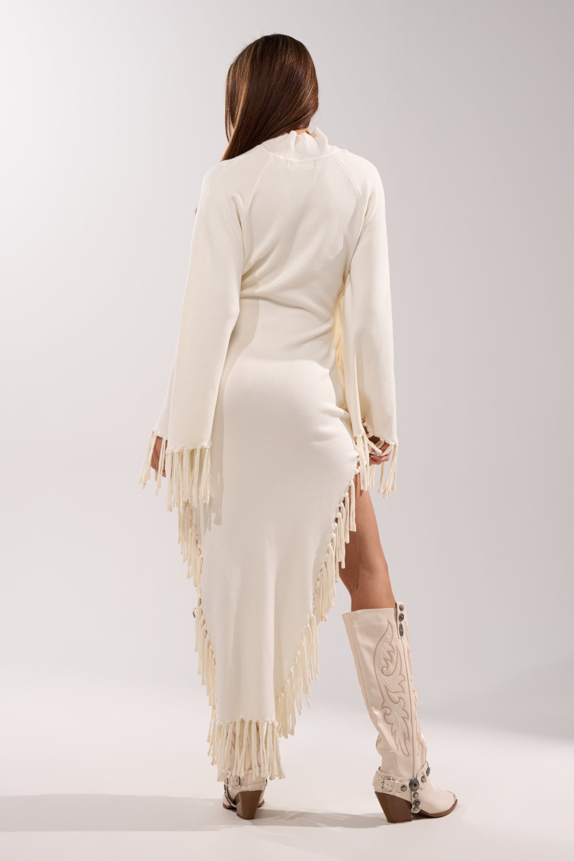 Model faces away in the SWEATER WEATHER KNIT MIDI DRESS, a cream knit midi with fringe trim and a high slit, styled with knee-high embroidered white boots. The background is plain and light-colored.