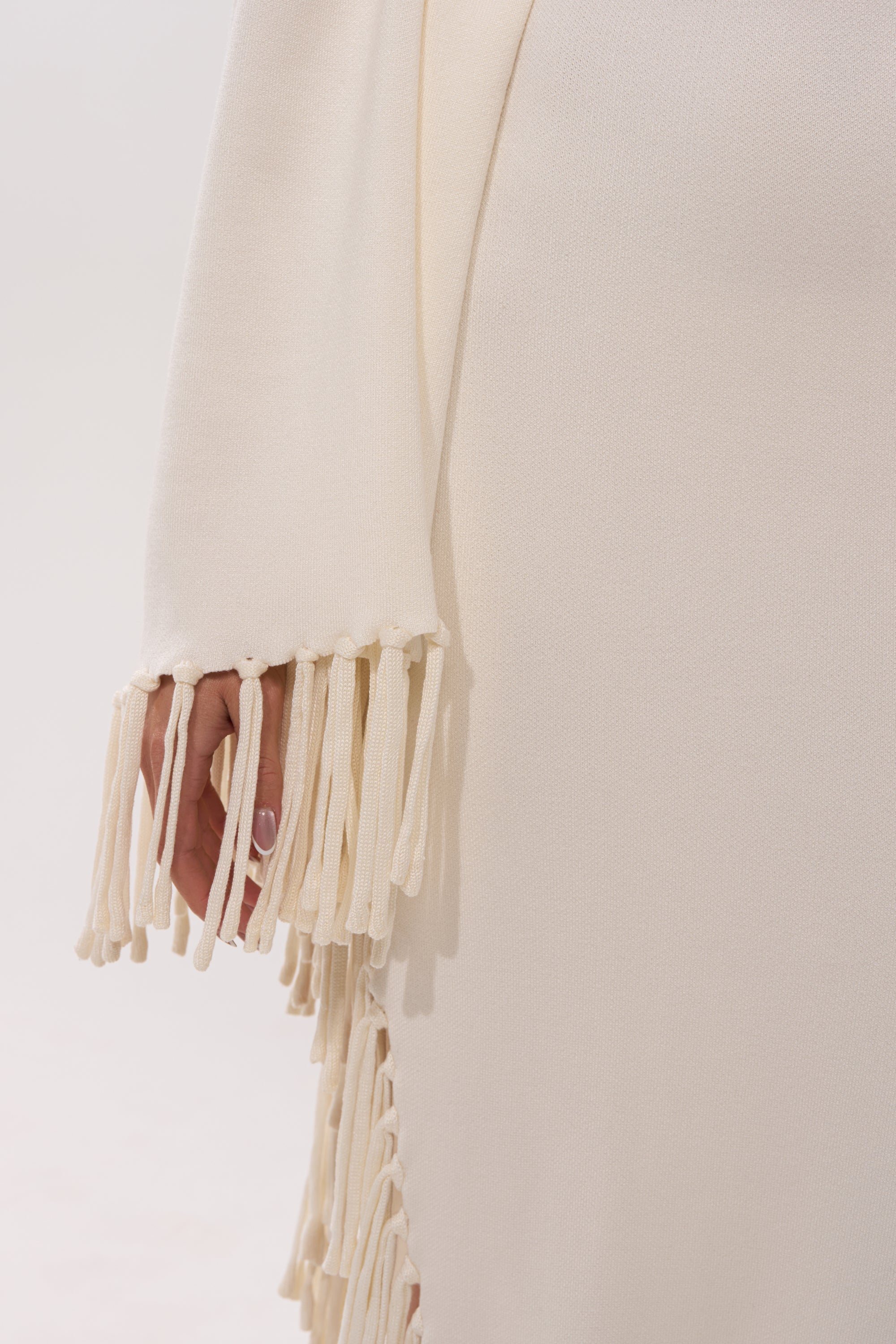 A close-up of a person in the SWEATER WEATHER KNIT MIDI DRESS, featuring cream knit fabric and long, textured fringe cascading from the cape sleeves. The fringe partly reveals their hand.