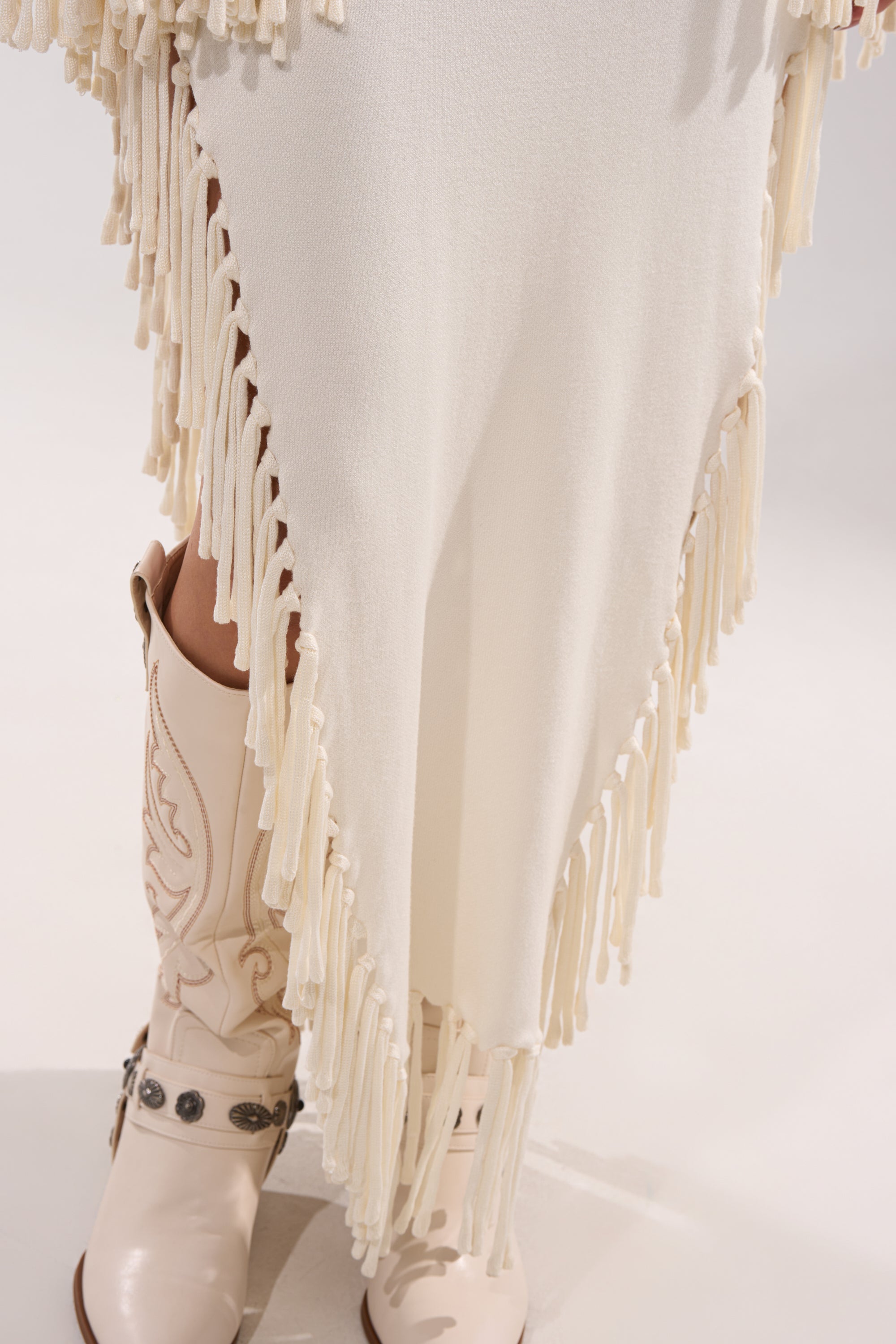 A close-up of a person wearing off-white cowboy boots with decorative stitching and the SWEATER WEATHER KNIT MIDI DRESS, which features long fringe trim along the edges.
