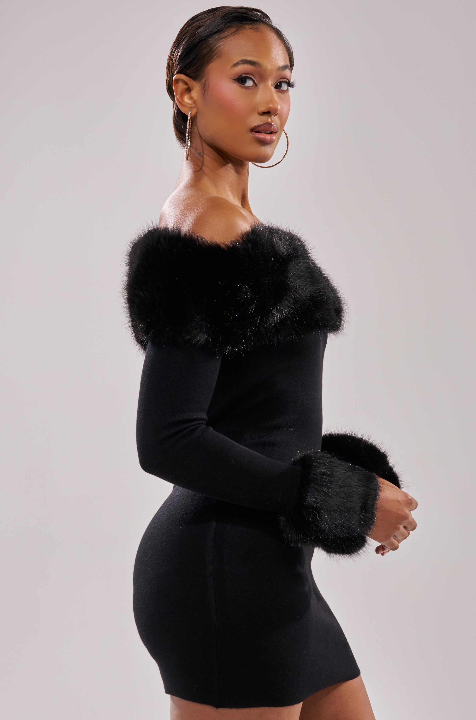 A woman poses against a plain light background, looking over her shoulder in the COMFY COZY FUR TRIM MINI DRESS IN BLACK—a fitted off-the-shoulder mini with luxe fur trim at the neckline and cuffs.