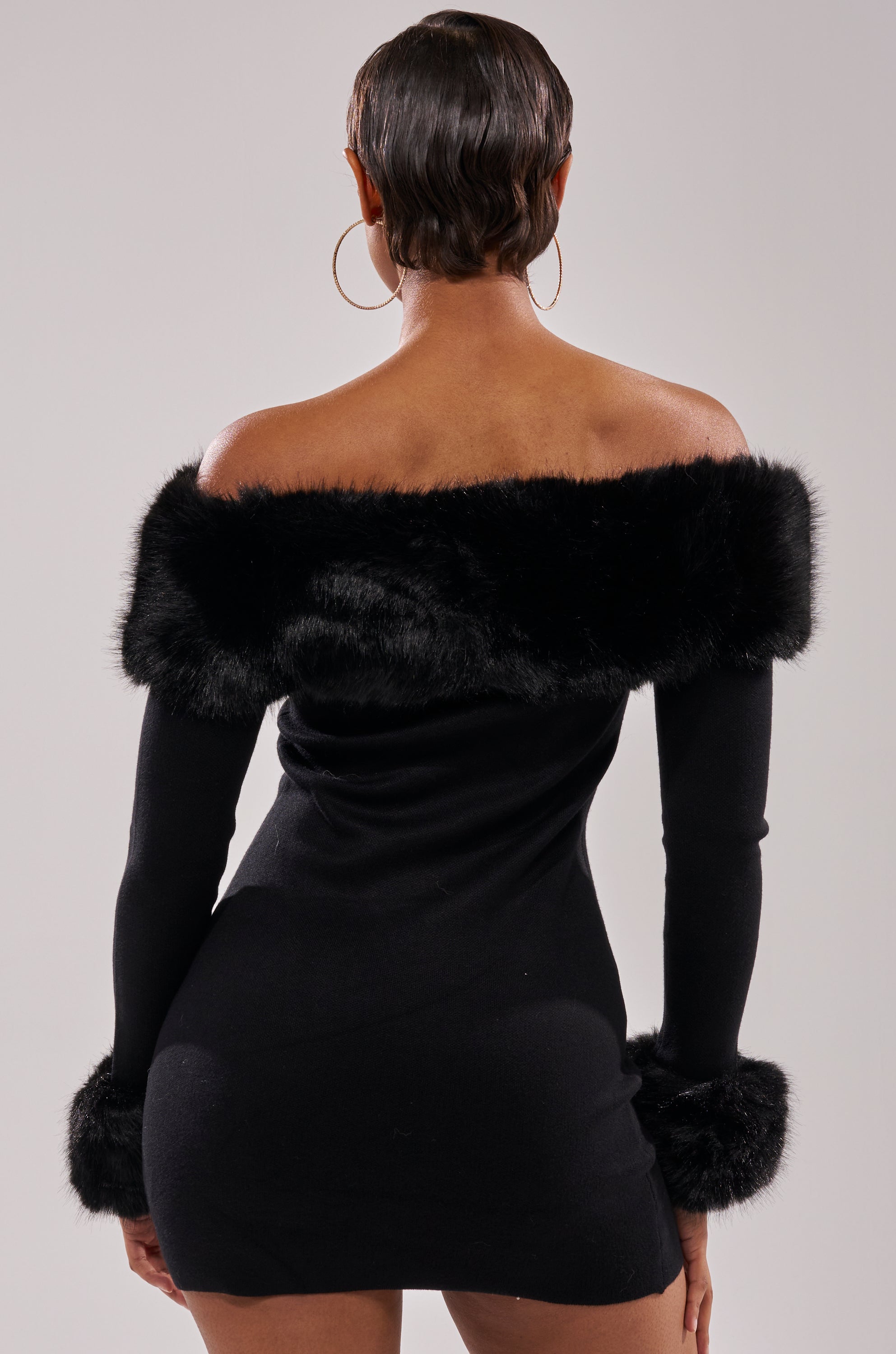A person with short hair and large hoop earrings models the COMFY COZY FUR TRIM MINI DRESS IN BLACK, standing with their back to the camera against a plain background.