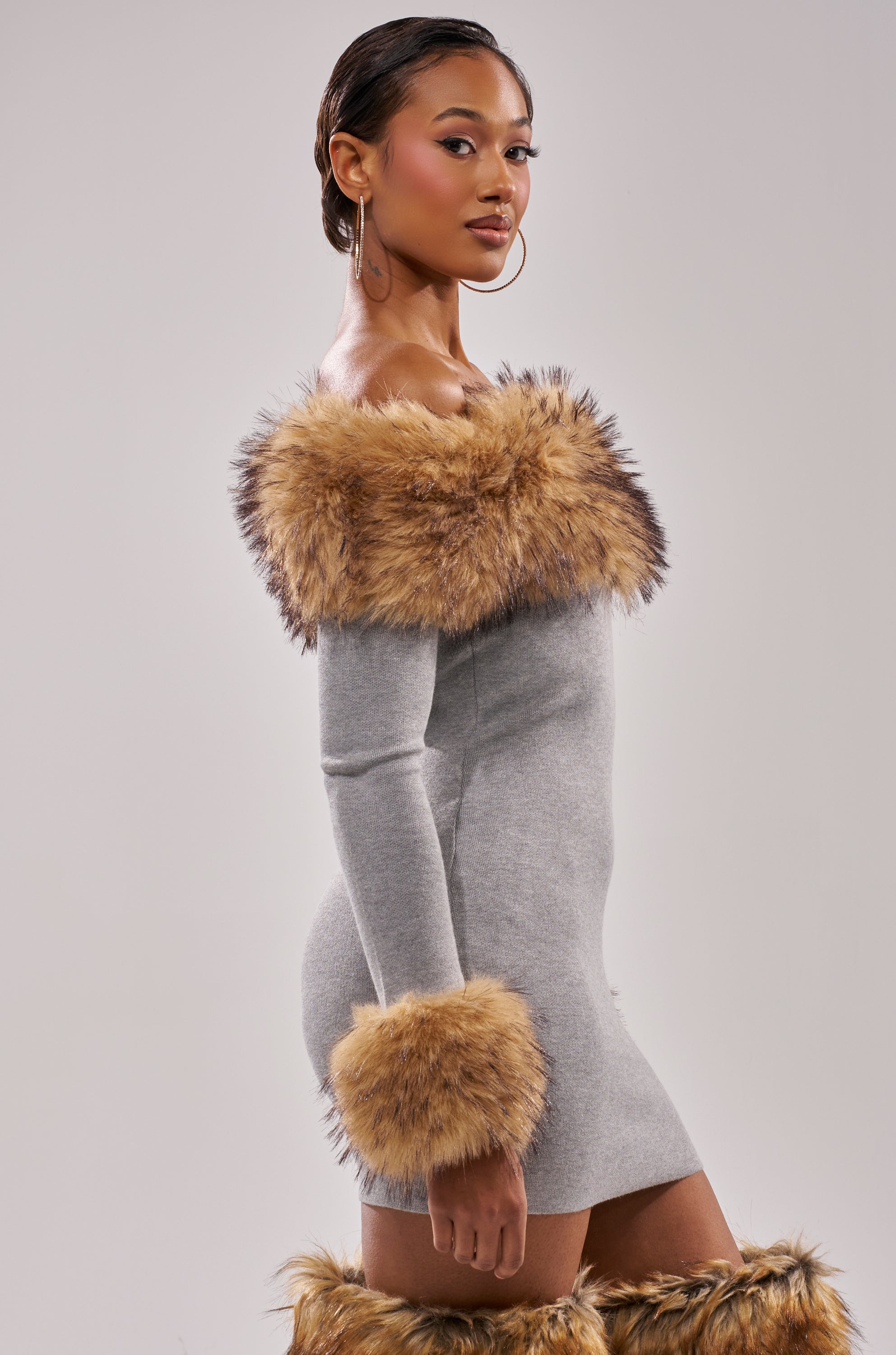 A woman models the COMFY COZY FUR TRIM MINI DRESS IN GREY, a fitted off-the-shoulder mini dress with grey faux fur trim on the neckline and cuffs. She pairs it with matching fur-trimmed boots, hoop earrings, and wears her hair styled back.