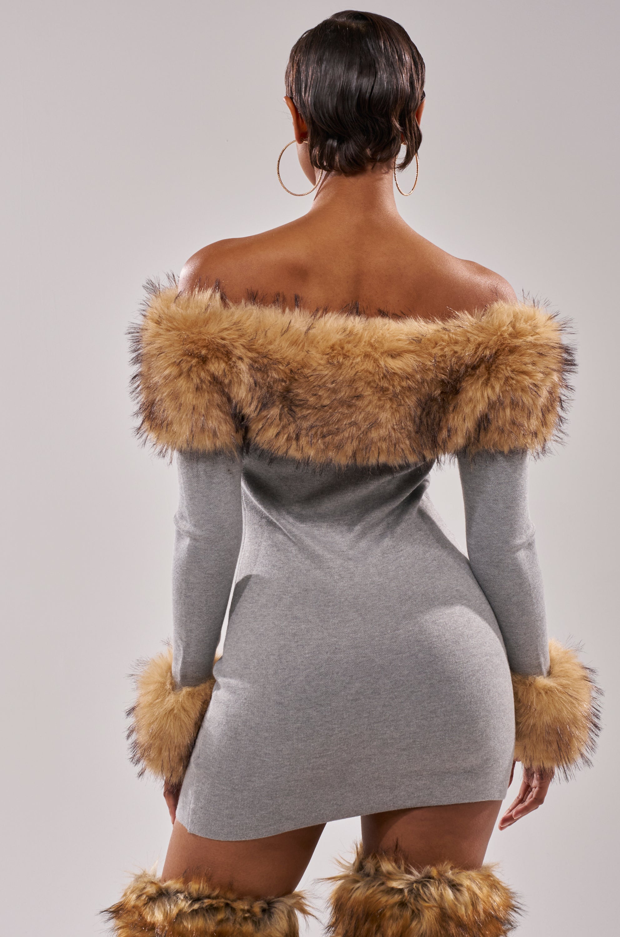 A woman with short hair, seen from behind, wears the COMFY COZY FUR TRIM MINI DRESS IN GREY featuring brown faux fur at the neckline and cuffs, plus matching boots and large hoop earrings, standing against a plain background.
