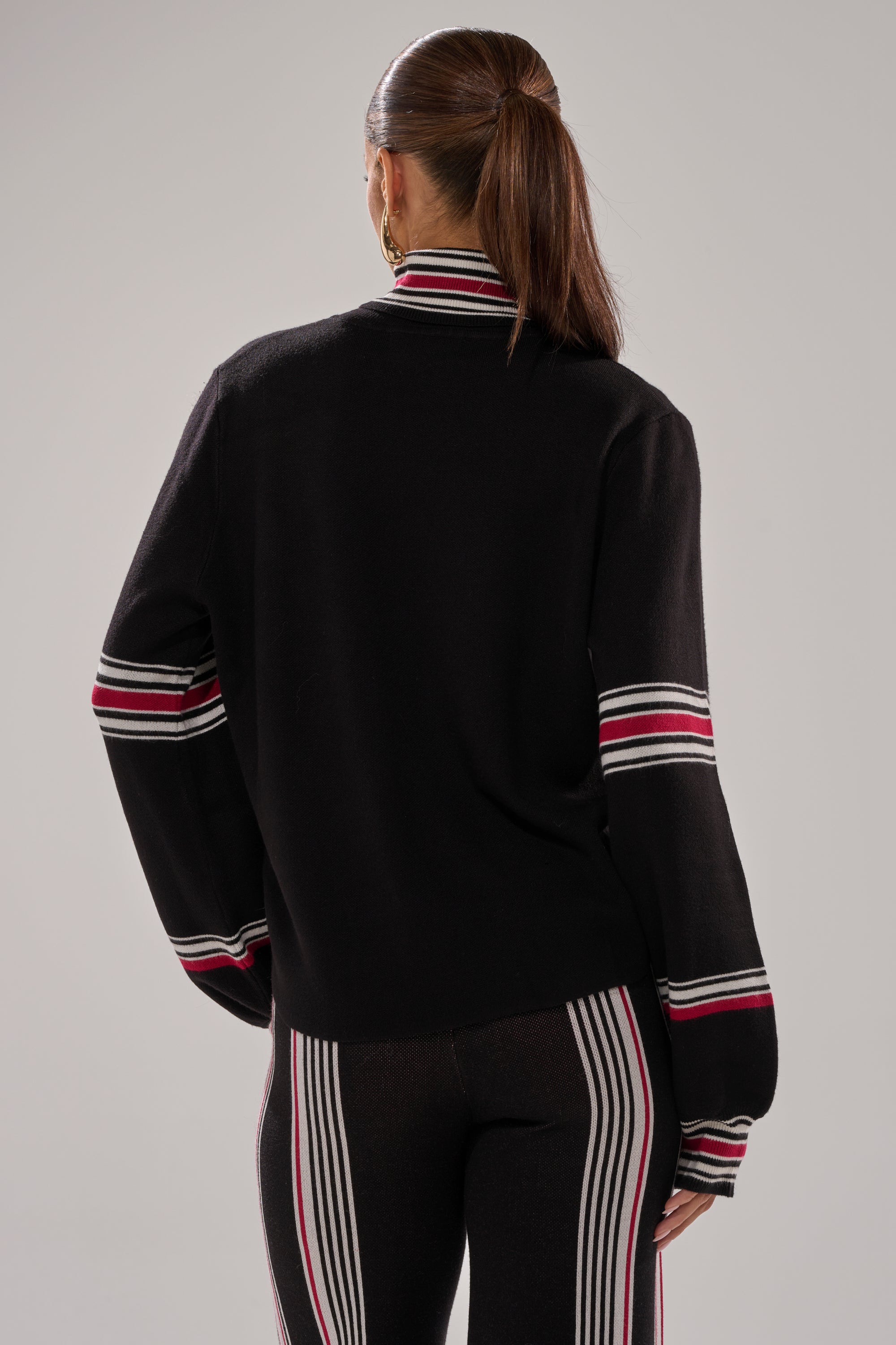 A woman with long brown hair in a ponytail is seen from behind, wearing the KENDALL TURTLE NECK SWEATER with red, white, and pink horizontal stripes and black pants, standing against a plain gray background.