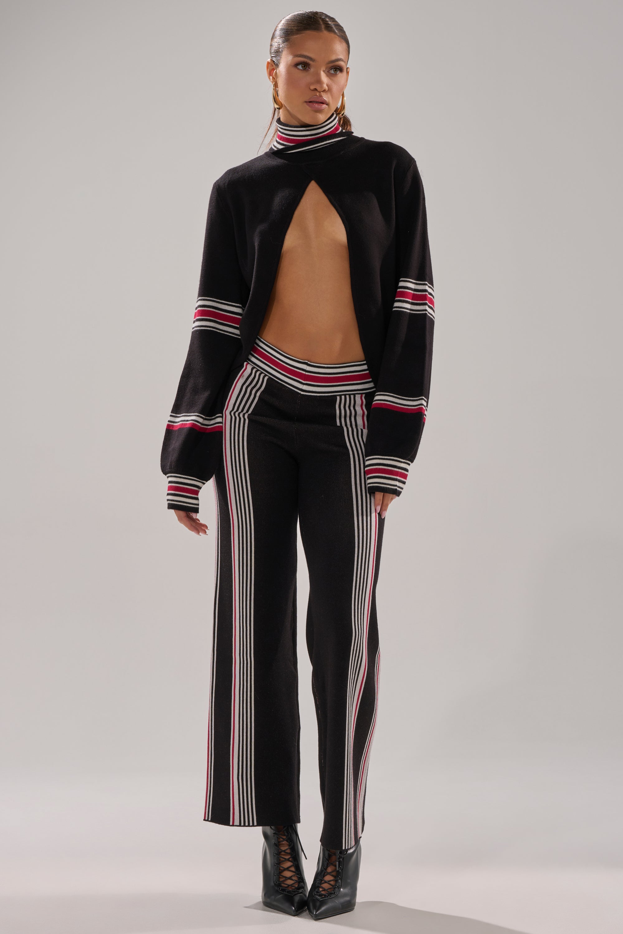 A woman wears the KENDALL TURTLE NECK SWEATER, a black knit with red, white, and pink stripes, styled open to reveal her midriff. She pairs it with matching striped pants and black heeled boots.