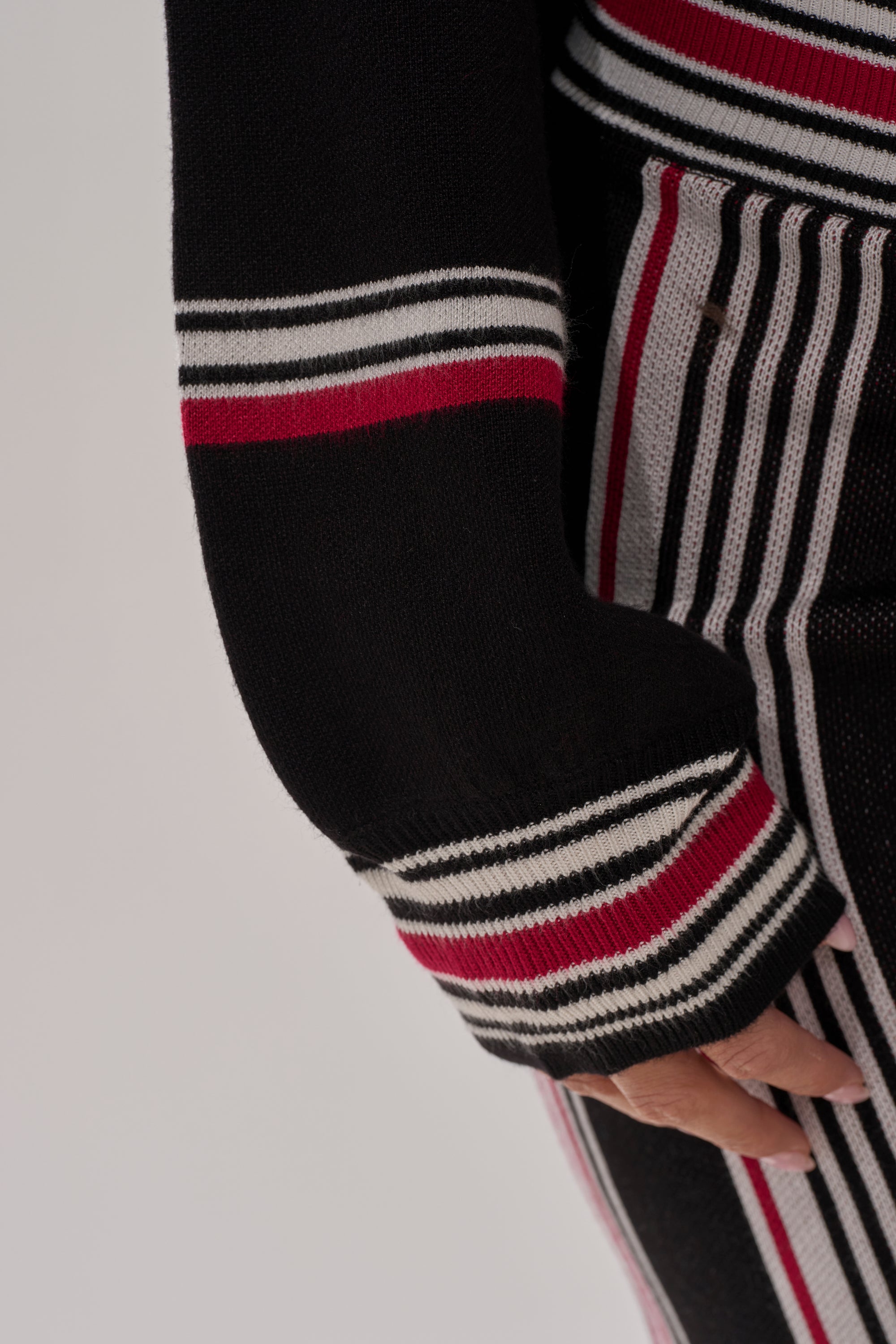 A close-up of a person’s arm and waist wearing the KENDALL TURTLE NECK SWEATER, featuring black fabric with horizontal red, white, and gray stripes on the sleeve. The hand is partially visible at the frame's edge.