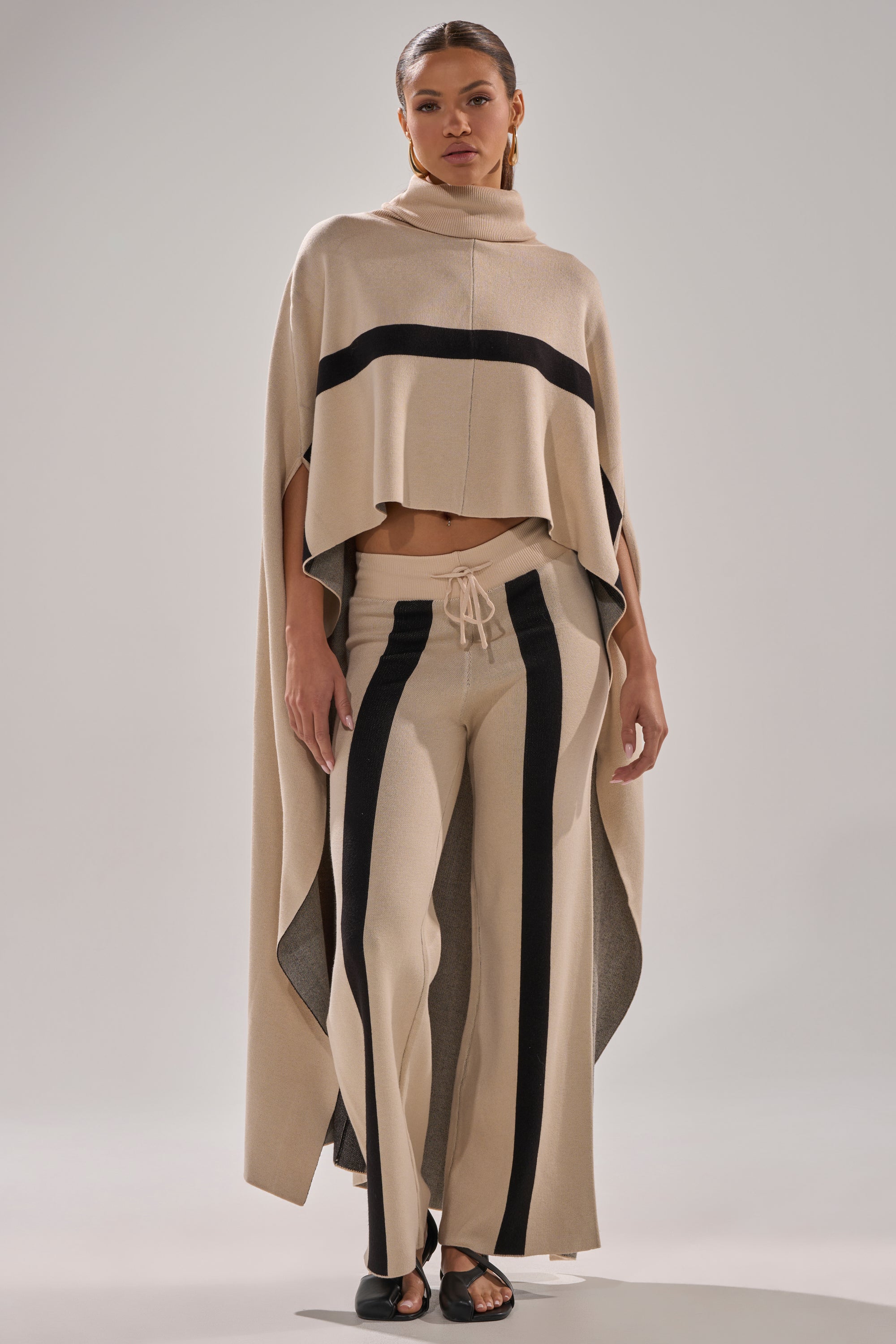 A woman wears the CLAUDIA SWEATER WIDE LEG PANT with vertical black stripes, paired with a matching beige poncho featuring black horizontal stripes and black open-toe shoes for a modern, relaxed look.