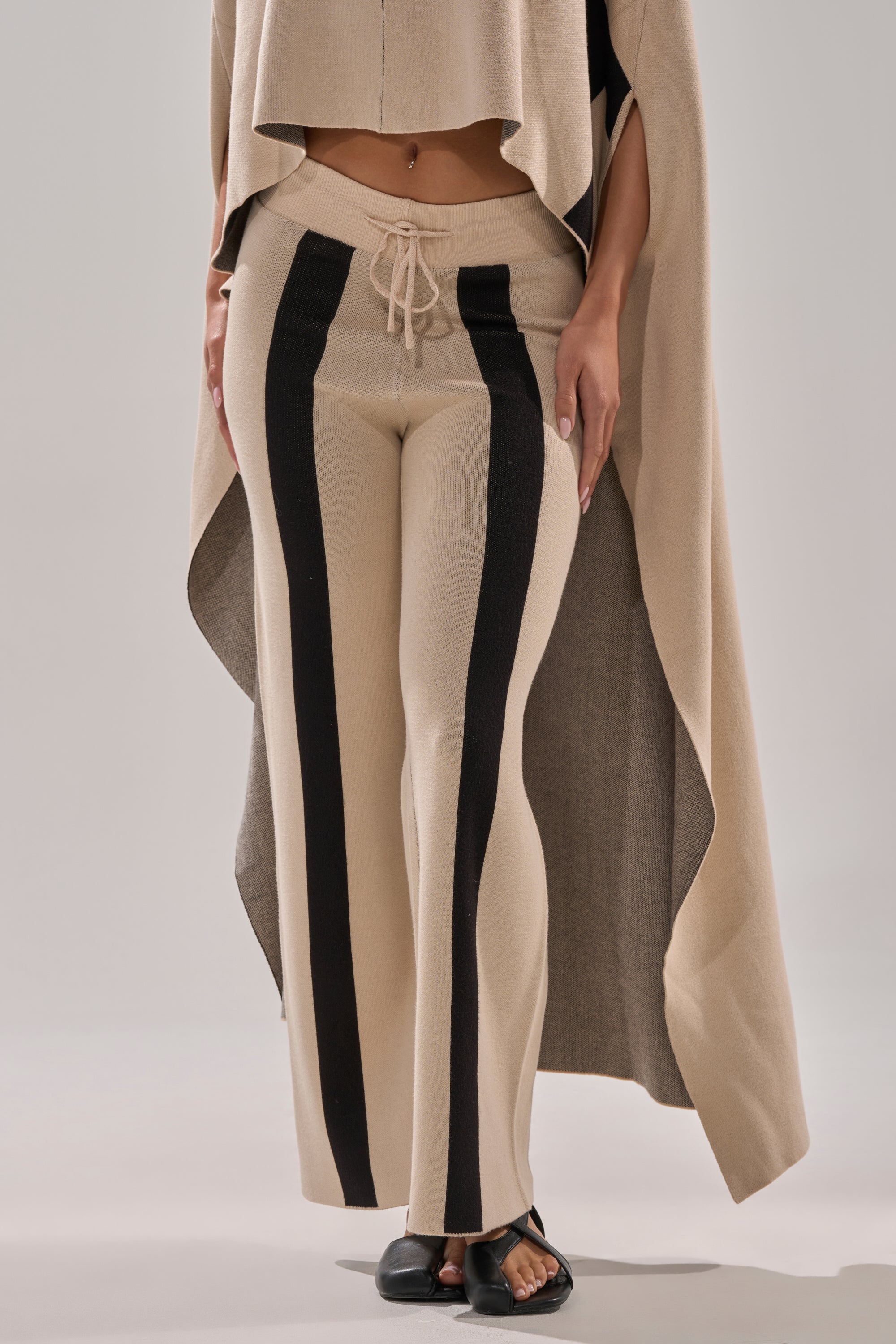 Wearing the CLAUDIA SWEATER WIDE LEG PANT featuring high rise, wide legs with bold black vertical stripes, paired with a matching beige poncho-style top, and standing barefoot on a light background.