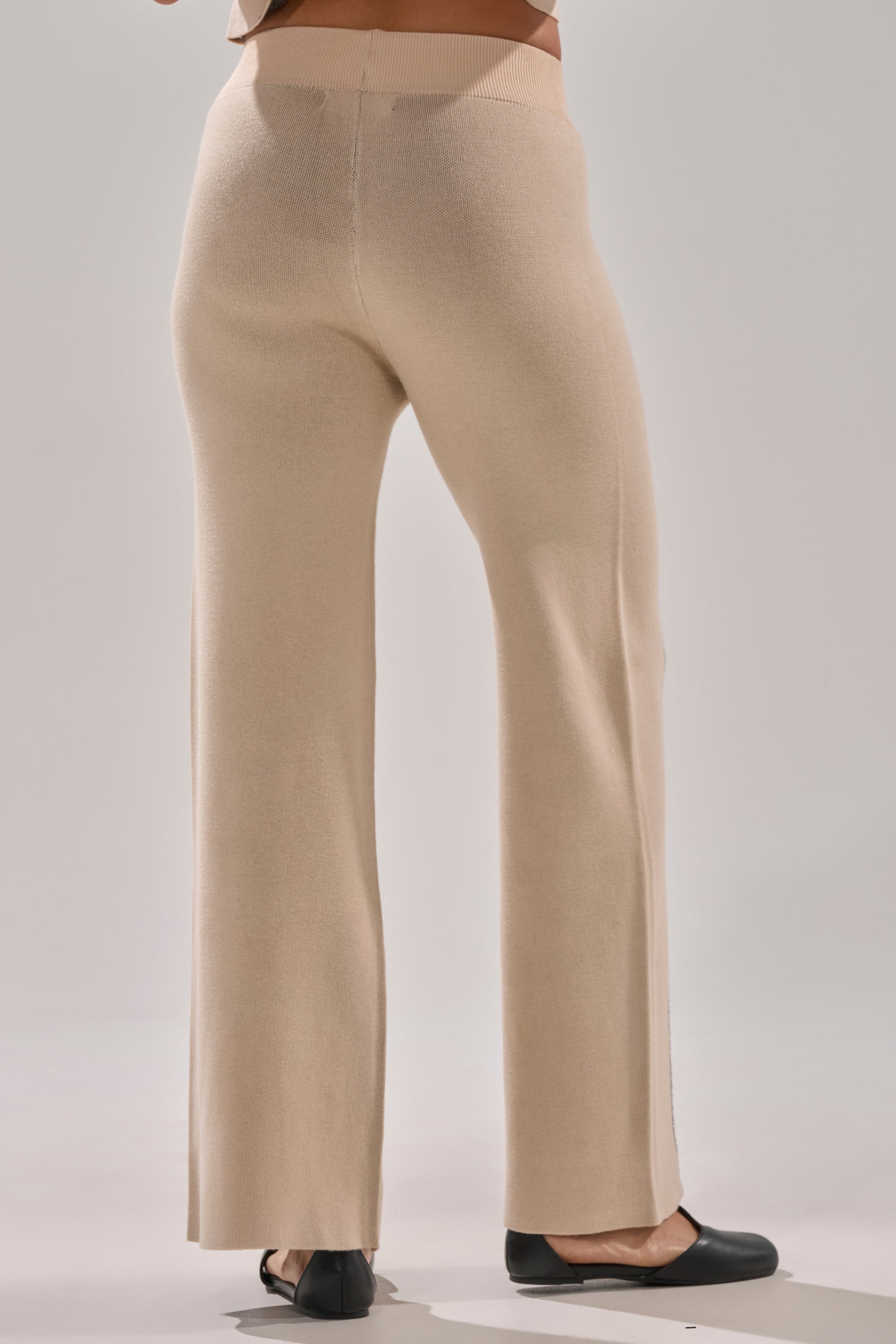 A person models the CLAUDIA SWEATER WIDE LEG PANT in beige with black ballet flats, standing waist down and facing away against a plain light gray background.
