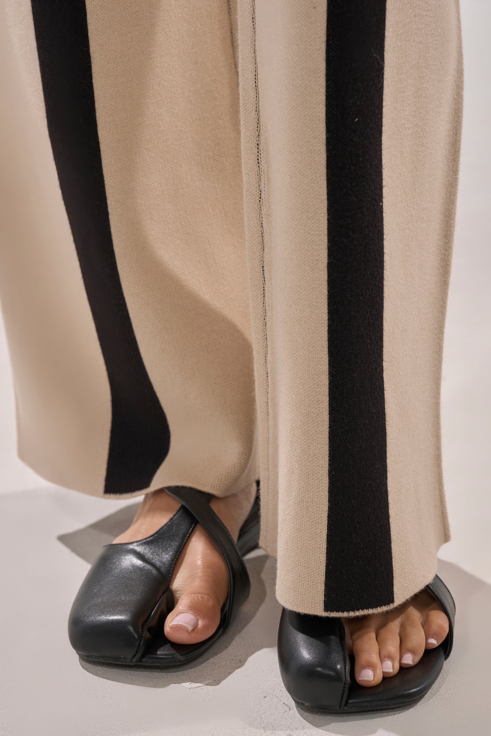 Close-up of a person’s feet in the CLAUDIA SWEATER WIDE LEG PANT, beige with vertical black stripes, paired with black open-toe sandals. The wide-leg pants reach the ground, partially covering the sandals and toes.