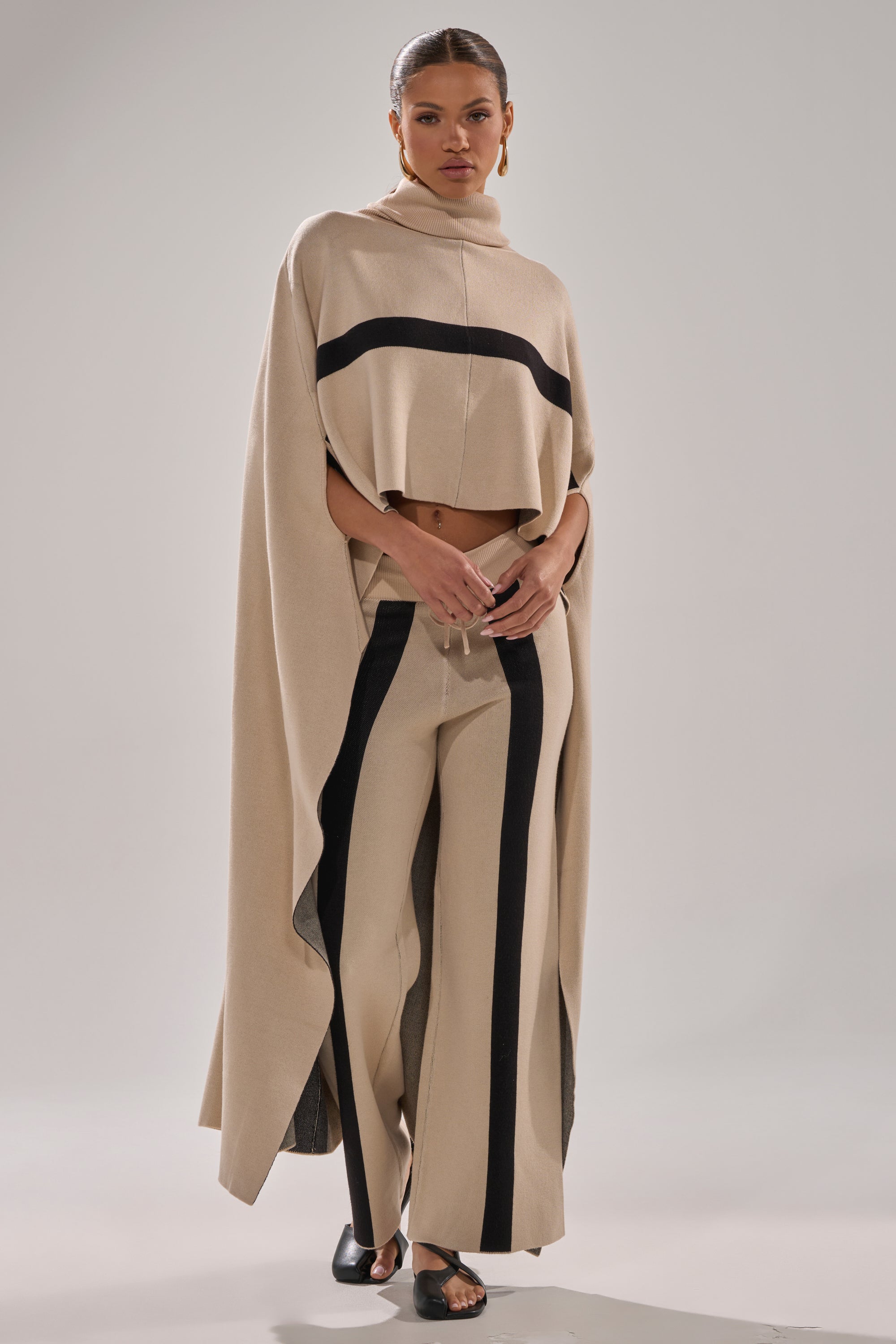 A woman poses against a plain background in the CLAUDIA TURTLENECK SWEATER with bold black stripes, paired with matching wide-leg pants, her hair pulled back, and finished with black open-toe heels.