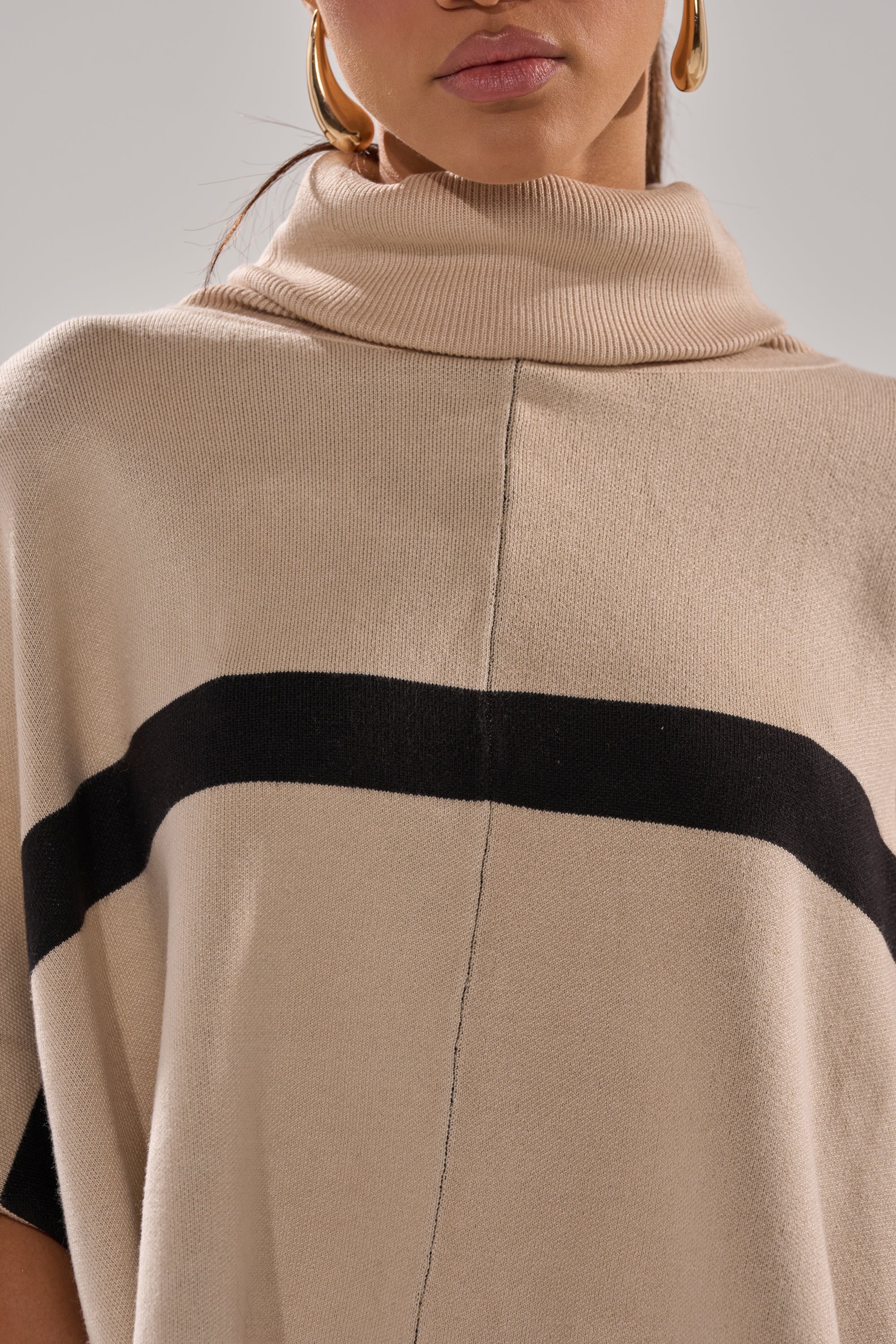 A woman models the CLAUDIA TURTLENECK SWEATER, featuring a beige hue with a black horizontal chest stripe. Only her lower face and upper torso are visible, and she pairs it with gold hoop earrings.